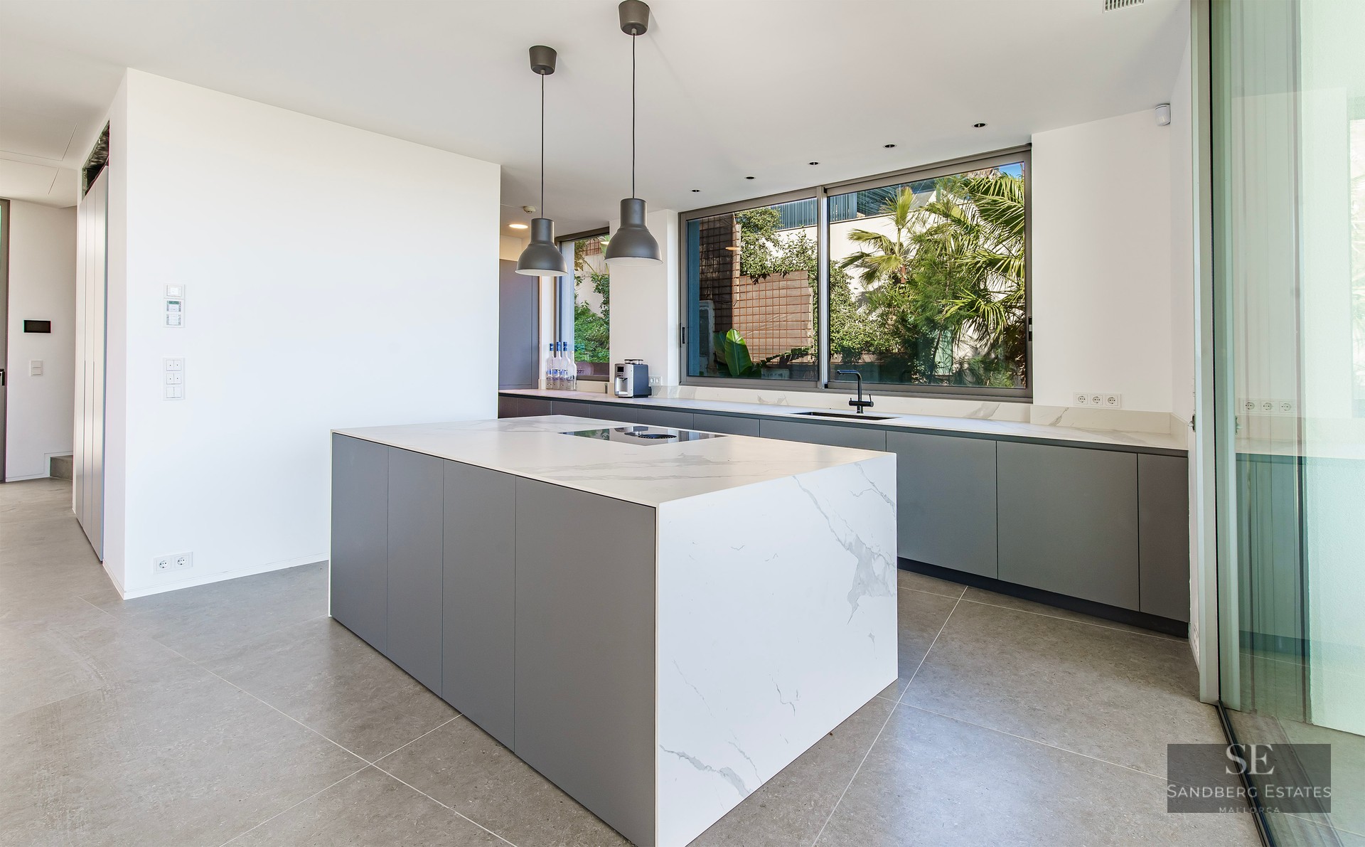 Modern kitchen with a large white marble island, grey cabinetry, and a window overlooking lush tropical plants.