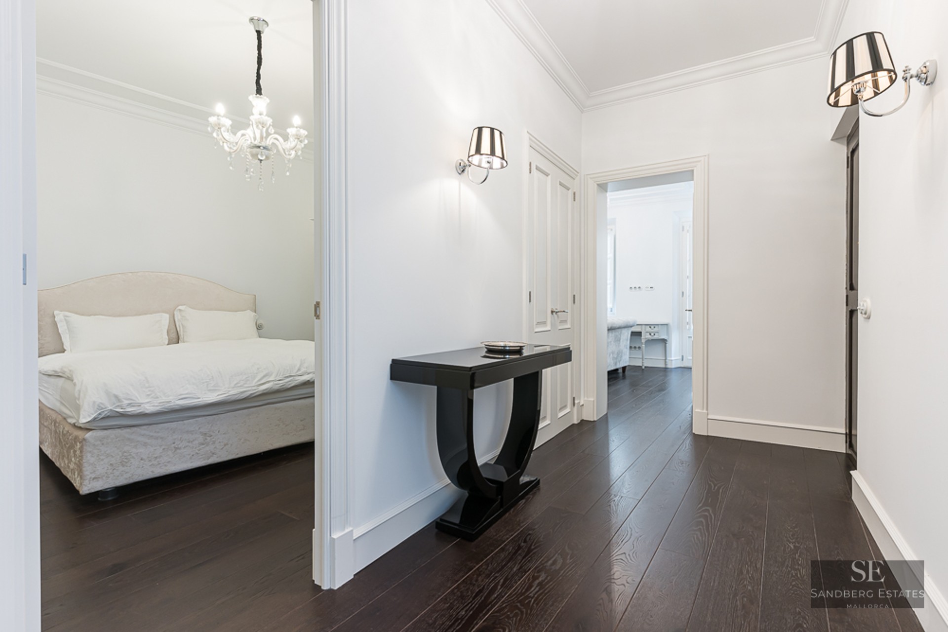 Bright hallway with dark wood floors, black console table, and view into a bedroom with a crystal chandelier.