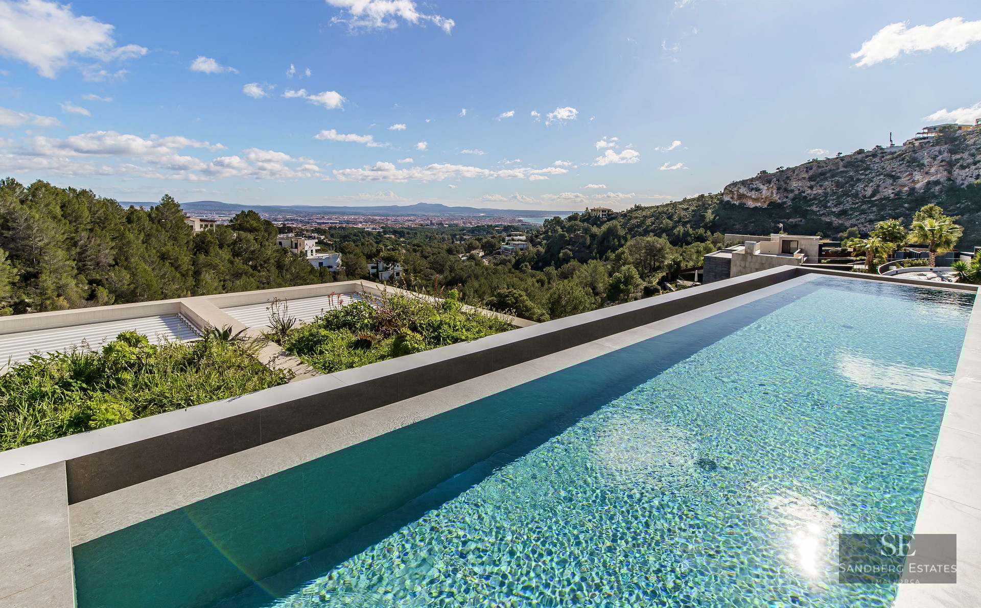 An infinity pool with clear blue water overlooking a green valley and distant city under a sunny sky.