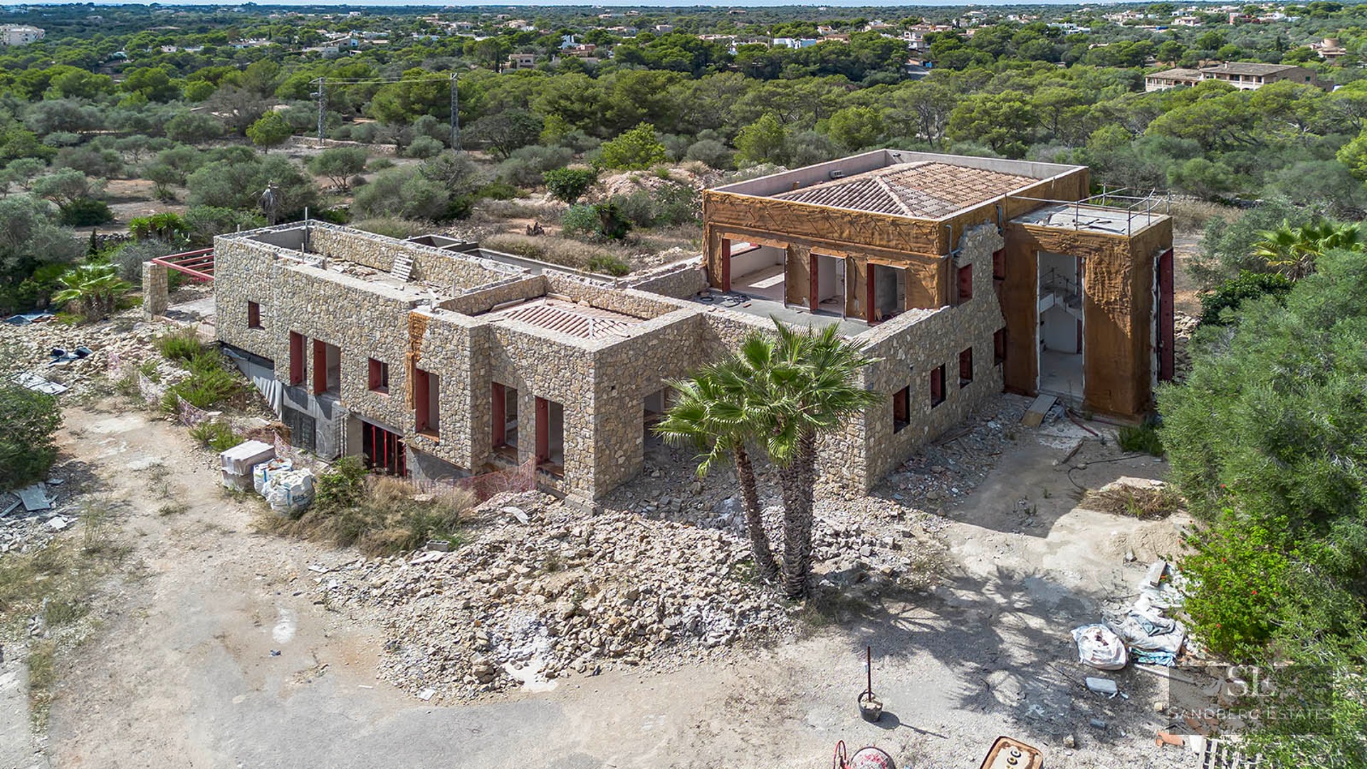 Elevated view of a large stone-walled luxury villa currently under construction surrounded by Mediterranean vegetation.