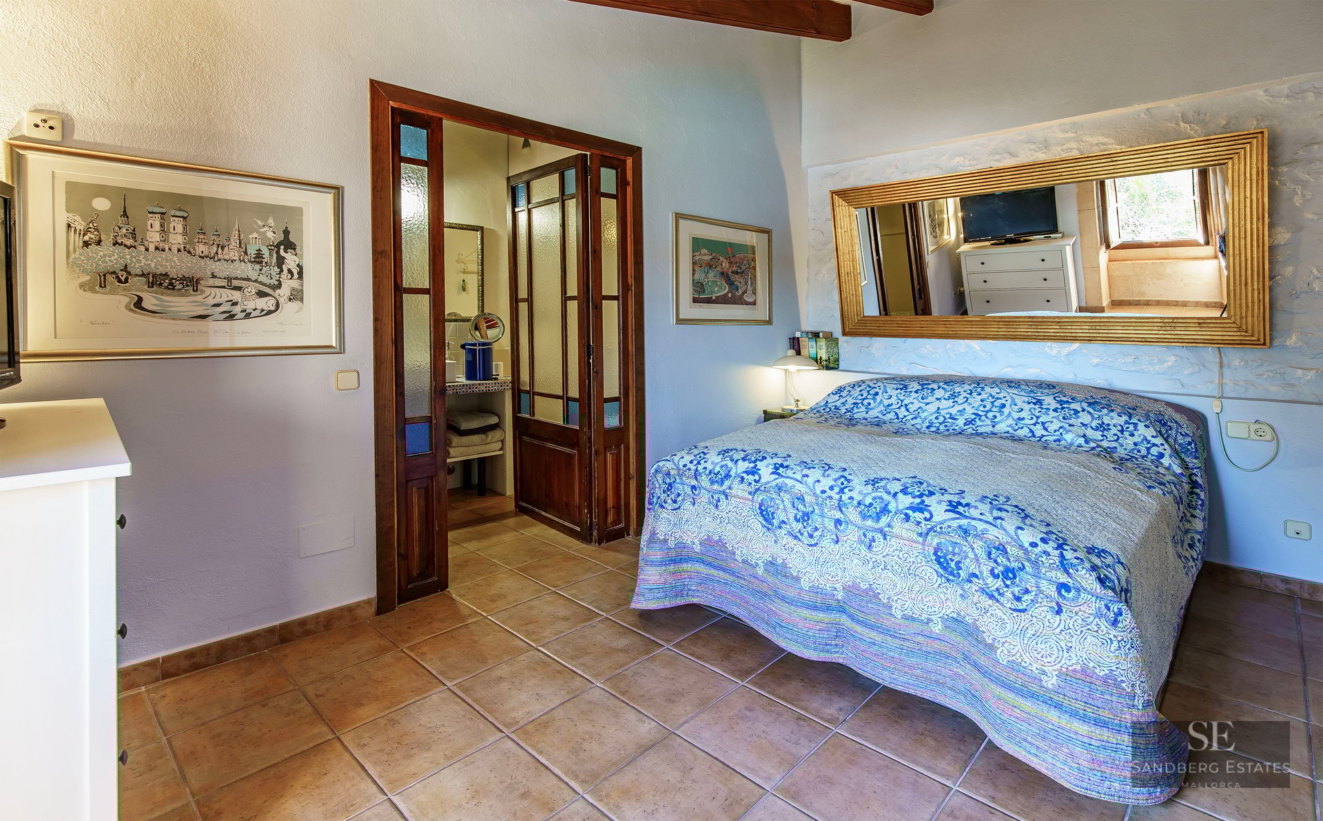 Bedroom with blue patterned bedspread, terracotta tile floor, large gold mirror, and dark wood doors to a bathroom.
