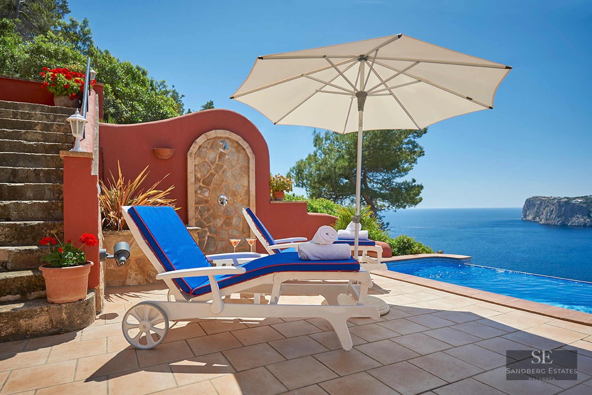 White sun loungers with blue cushions beside an infinity pool overlooking the Mediterranean Sea and cliffs.