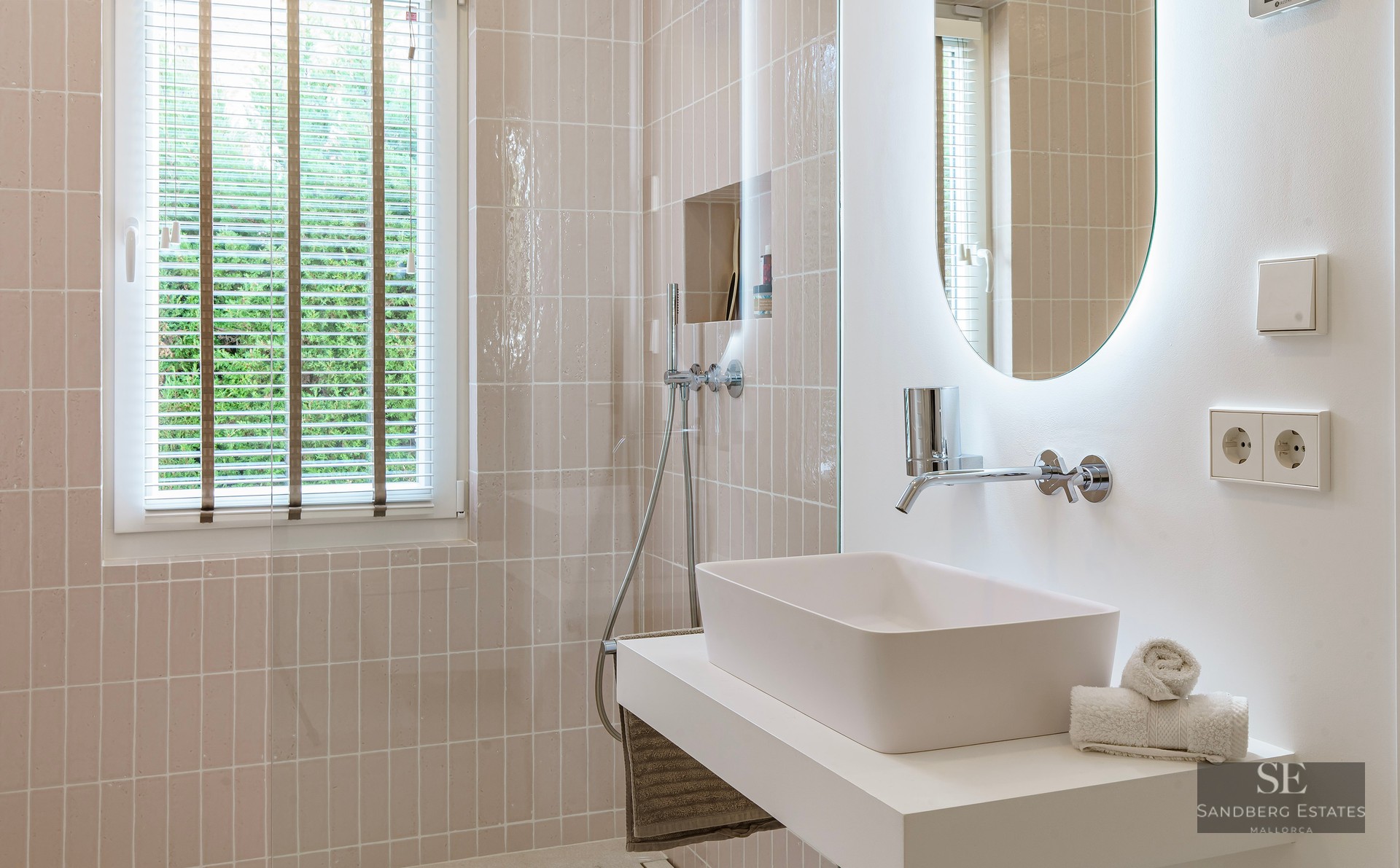 A clean, modern bathroom featuring a white vessel sink, backlit oval mirror, and soft beige vertical tiling.