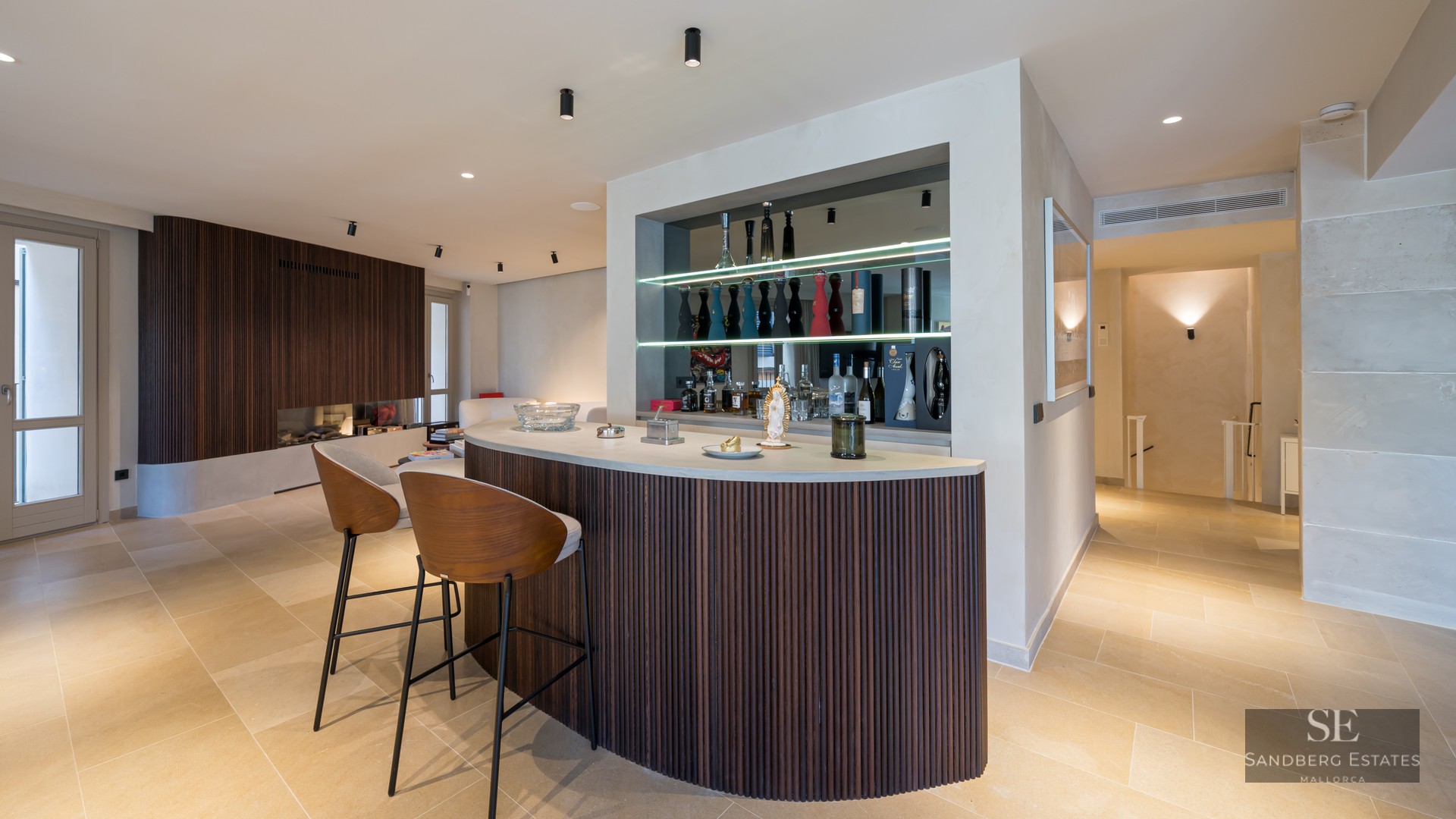 Curved home bar with dark wood slats, two designer stools, and backlit shelves with premium liquor bottles.