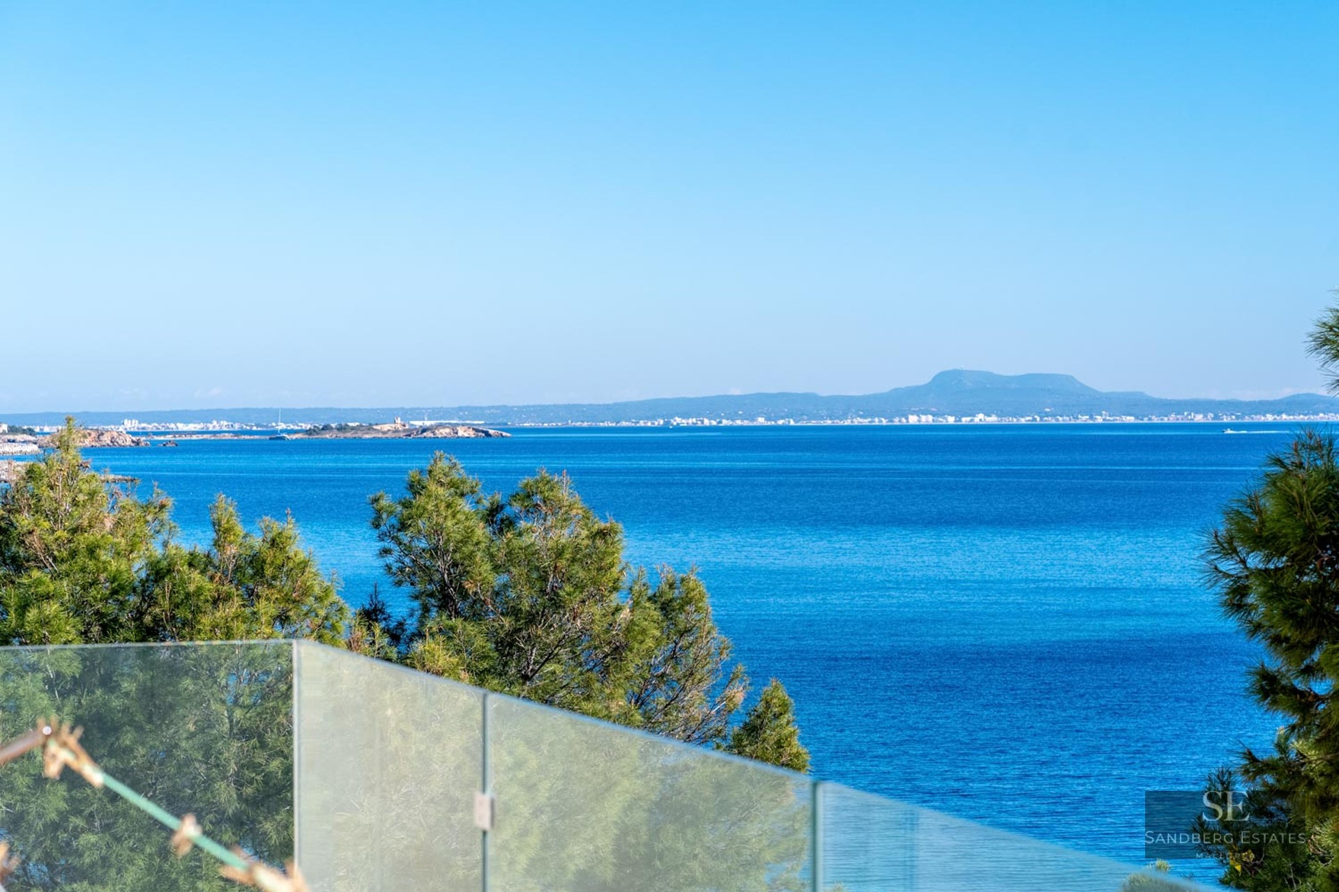 View of the deep blue Mediterranean sea and distant coastline from a terrace with a glass railing and pine trees.