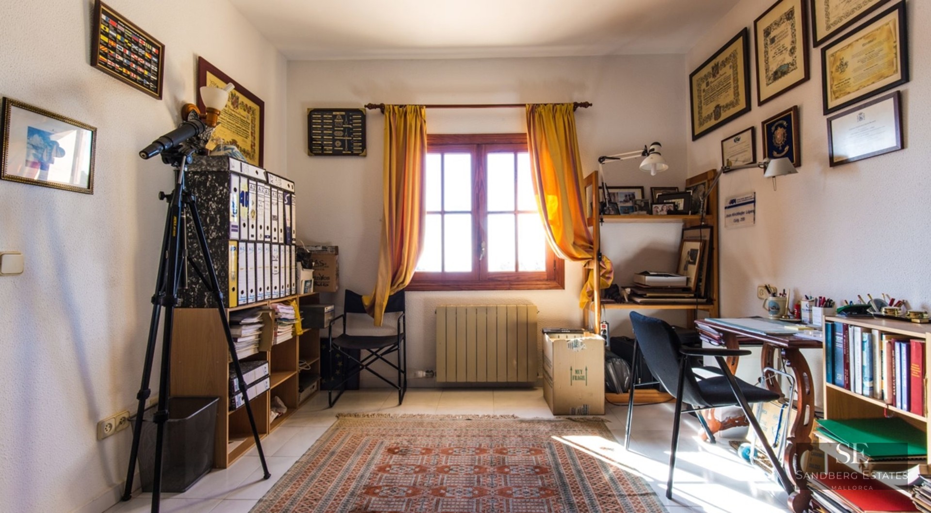 A home office with a wooden desk, telescope, bookshelves, yellow curtains, and certificates on the walls.