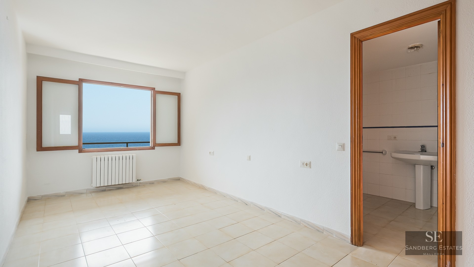 Empty white room with tile floors, a wooden-framed window looking out to the blue sea, and an open door to a bathroom.