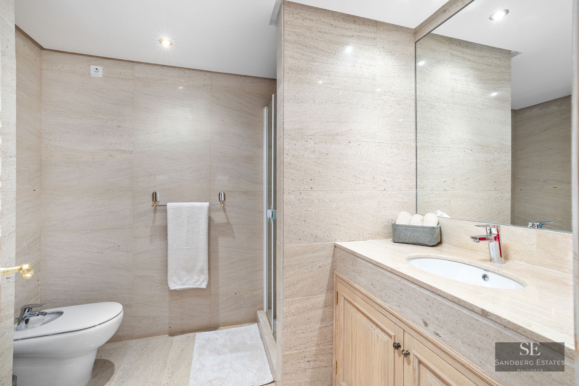 Salle de bain moderne avec murs en pierre beige, bidet blanc, meuble en bois avec lavabo et grand miroir.