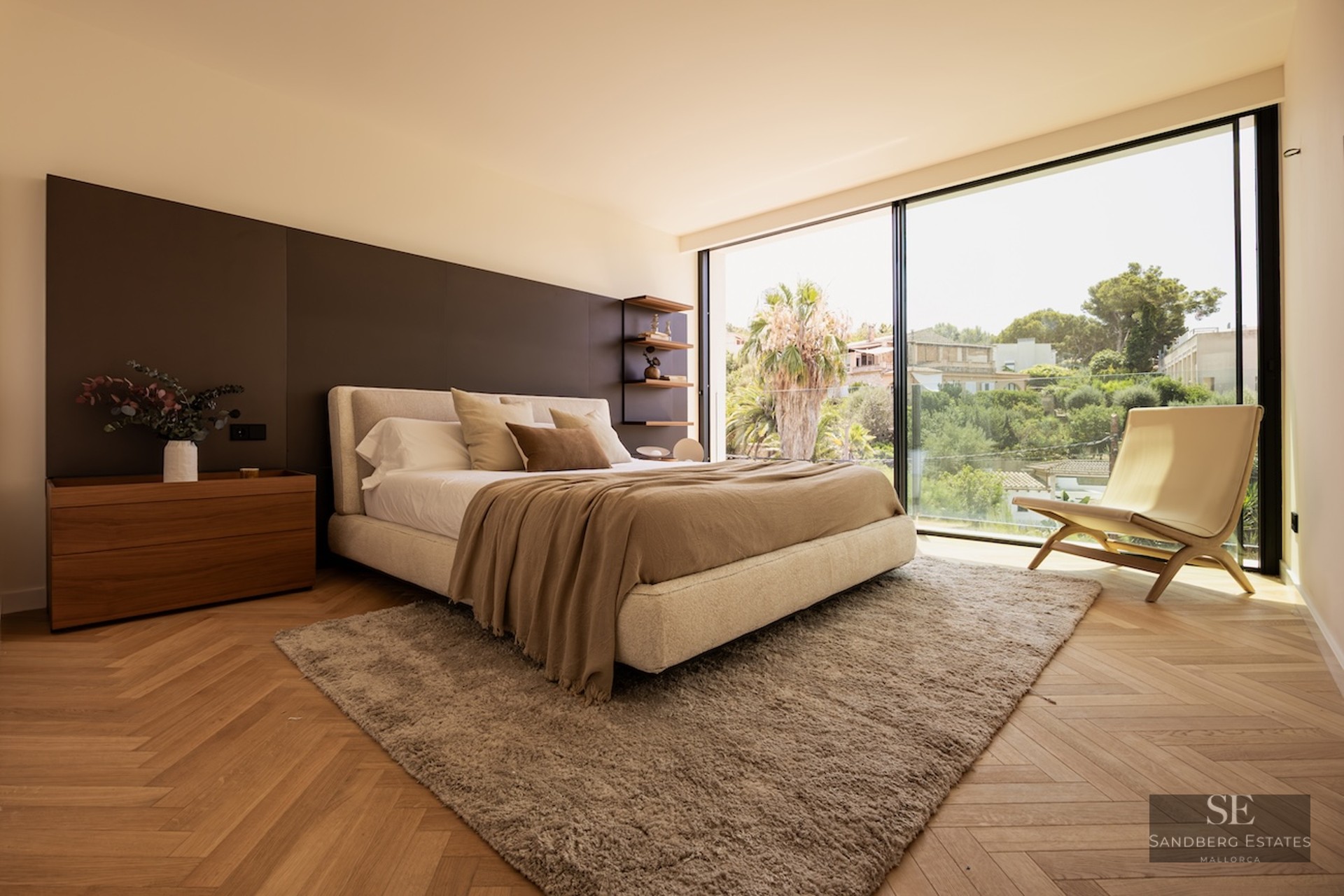 Bright master bedroom with herringbone wood floors, a large bed, and floor-to-ceiling windows overlooking lush greenery.