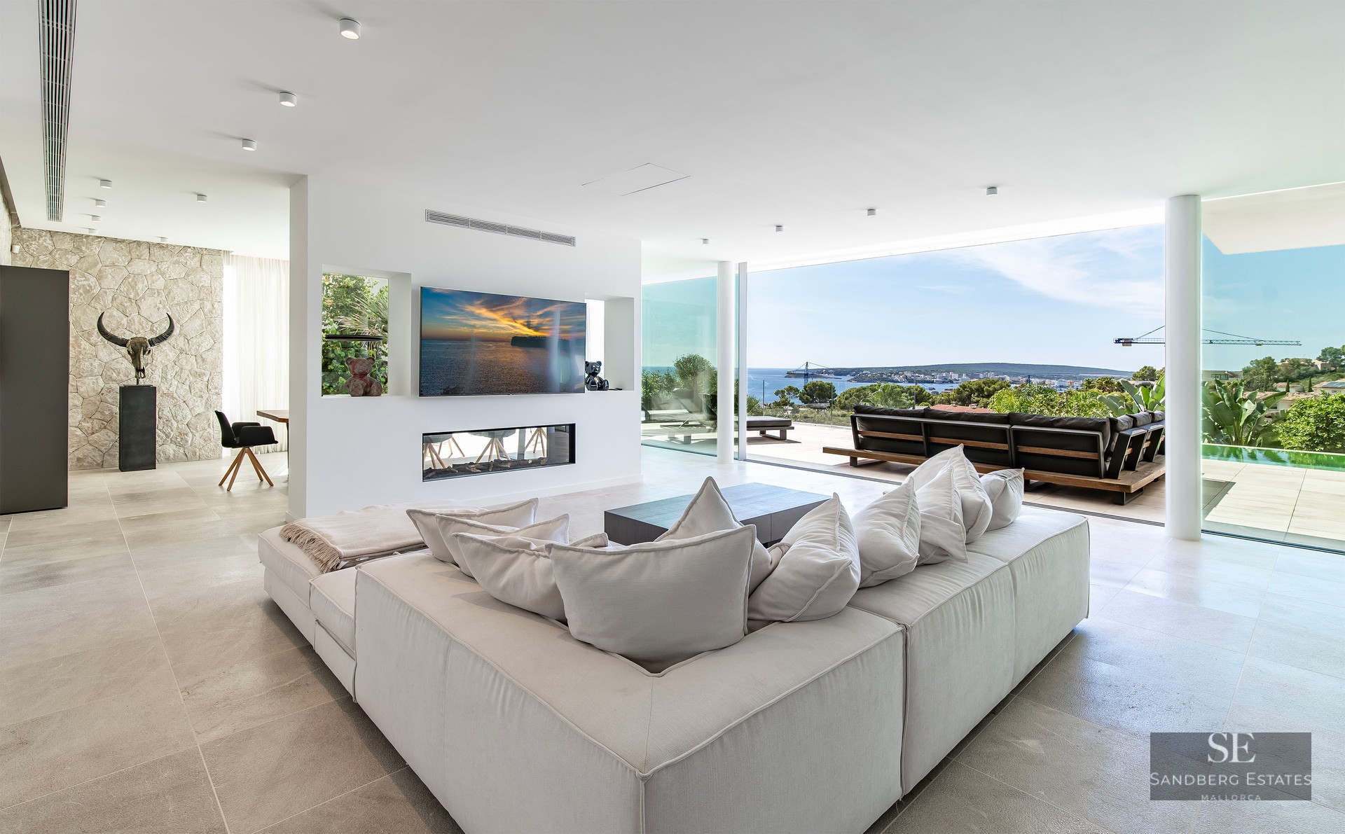 Spacious modern living room with large white sectional sofa, integrated fireplace, and floor-to-ceiling windows with sea views.