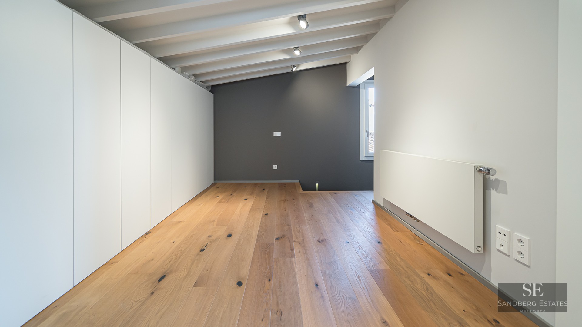 Empty bedroom featuring light wood floors, a white beamed ceiling, gray accent wall, and large white wardrobes.