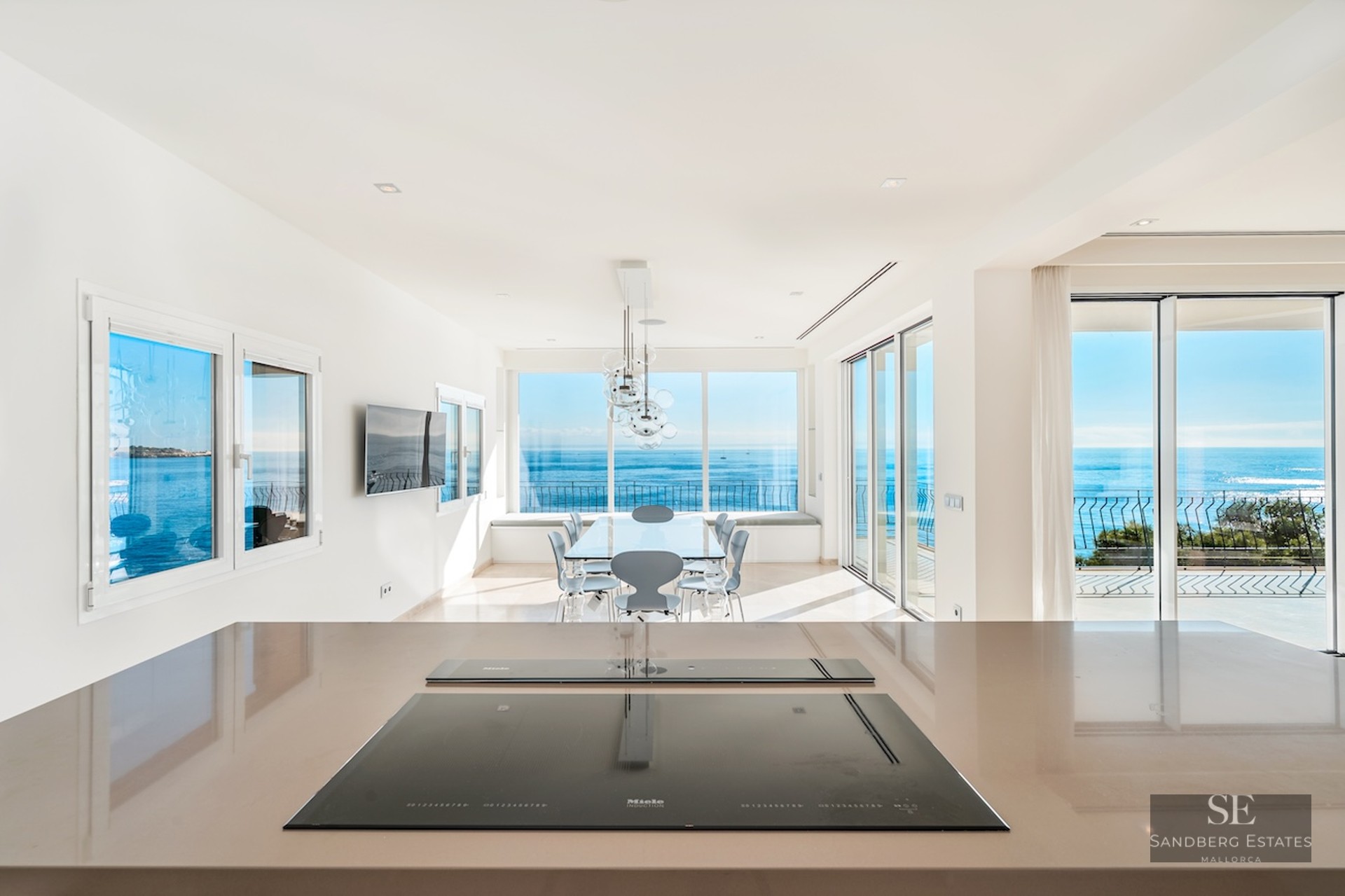 Close-up of a sleek kitchen counter with an induction hob, looking out towards a dining area and a wide blue sea view.