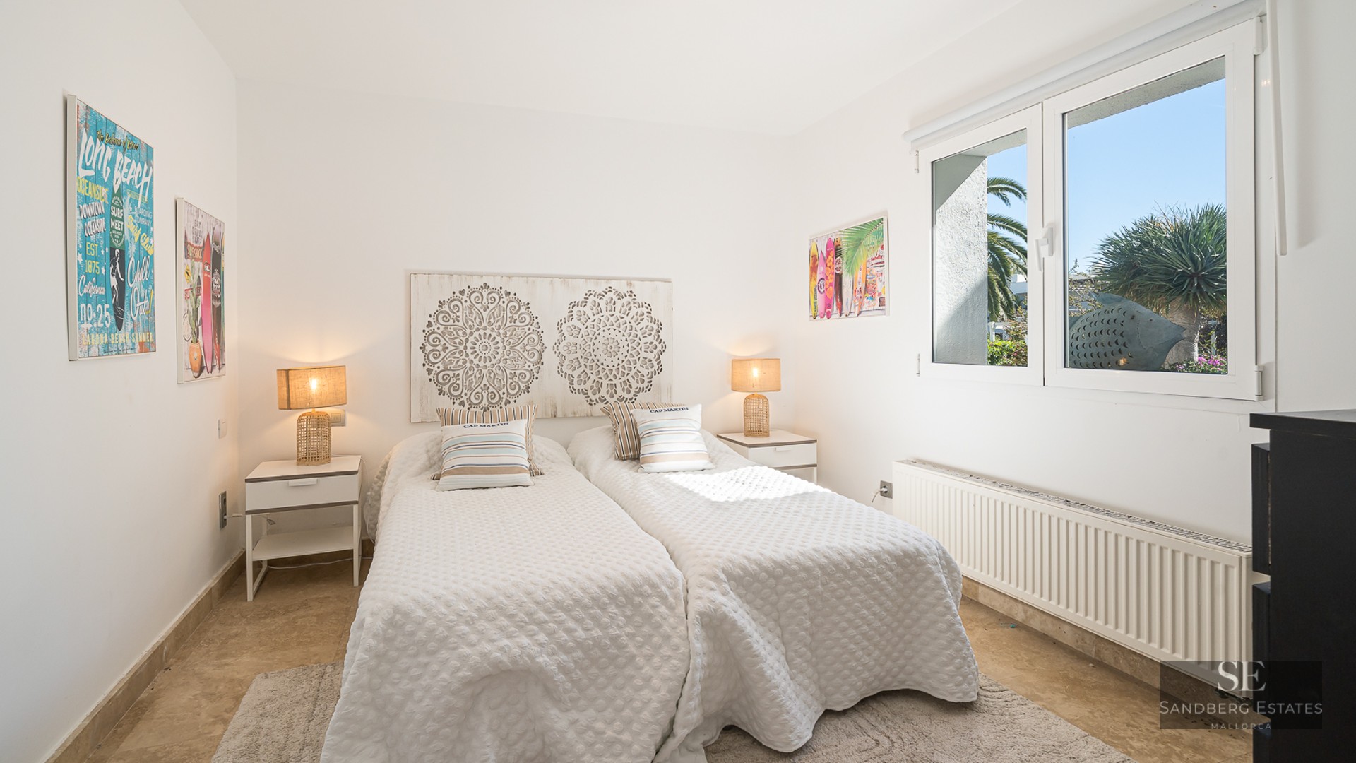 White room with two twin beds, carved wooden headboard, wicker lamps, and a window showing garden views.