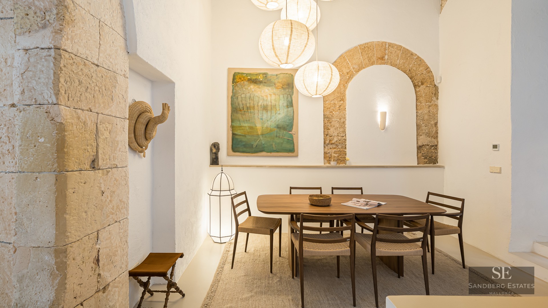 A wooden dining table with six chairs under paper globe lamps, set against white walls with a rustic stone arch.