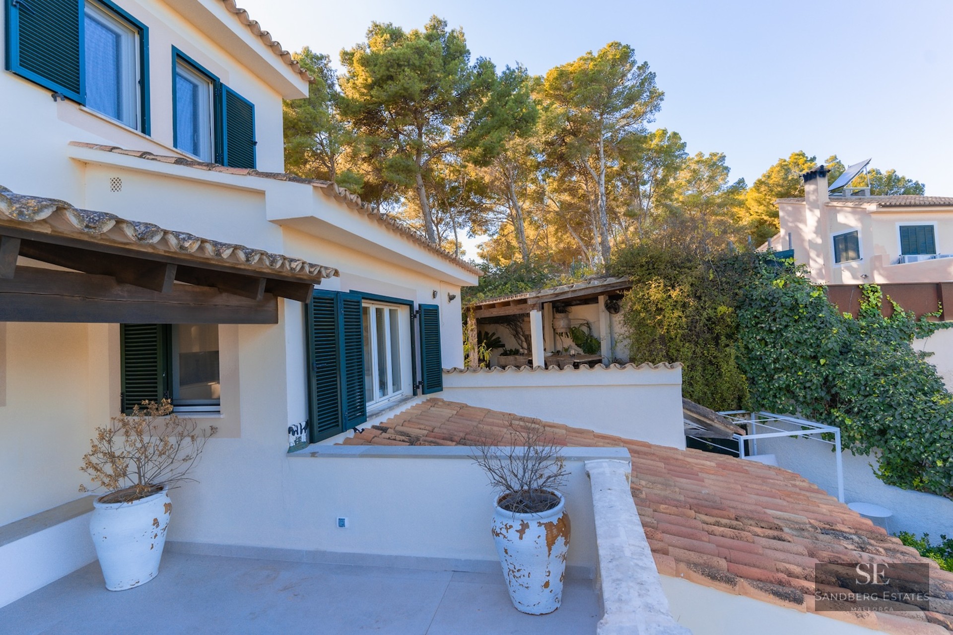 White Mediterranean villa exterior with terracotta roof tiles, dark green shutters, and large rustic white planters.