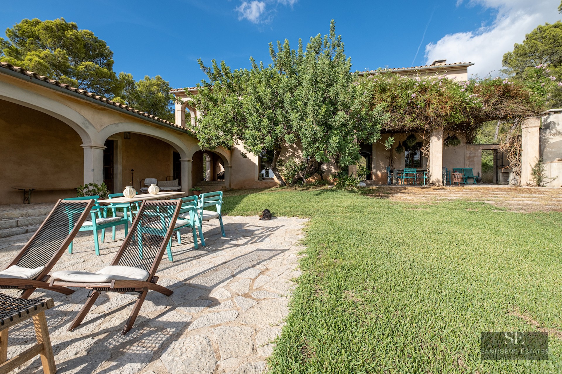A sunny garden with a stone patio, lawn, teal chairs, wooden loungers, and a rustic villa with arched porches.