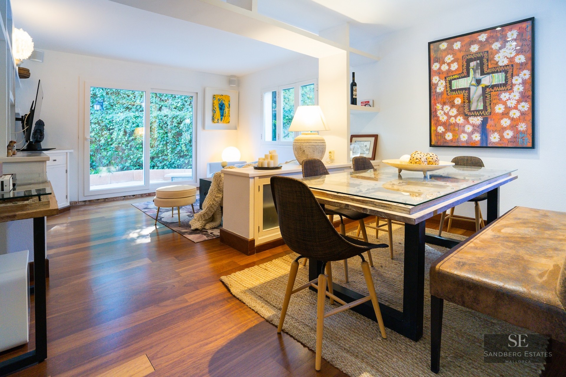Bright dining area with a glass-topped table, designer chairs, and large artwork looking toward a garden view.