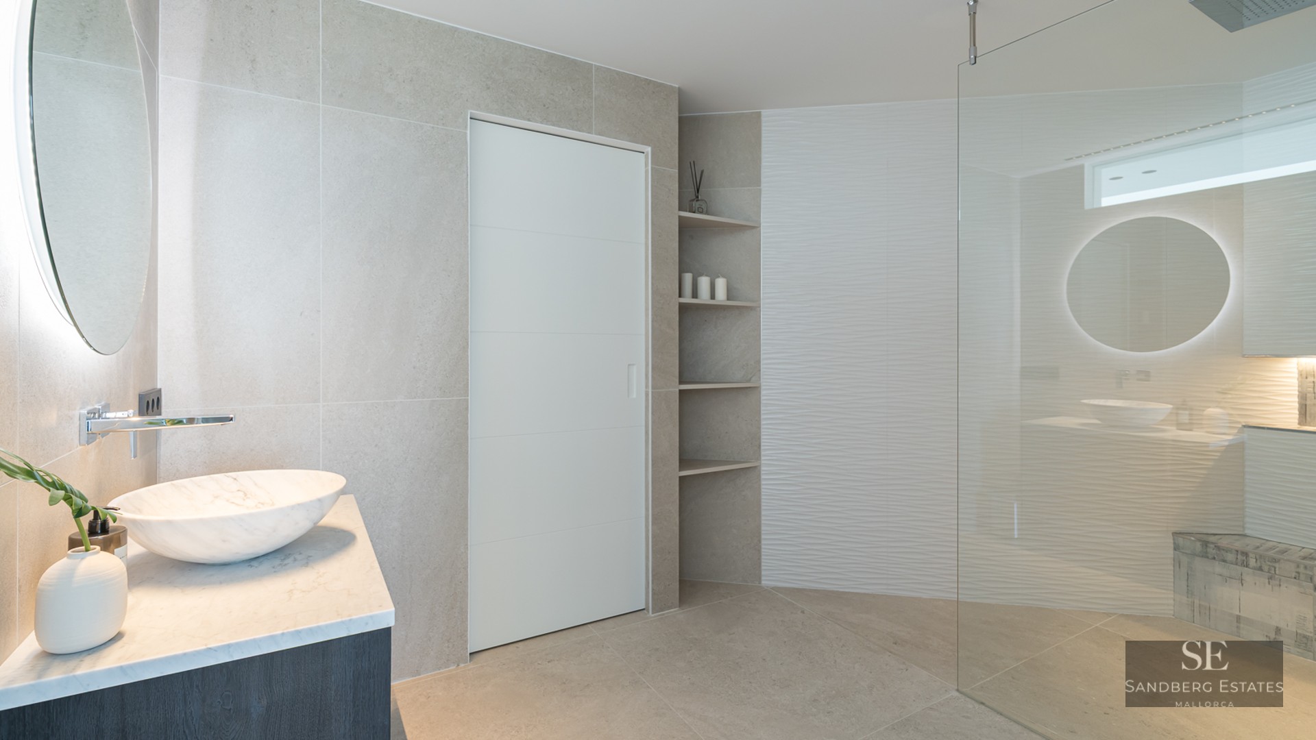 Modern bathroom featuring a marble vessel sink, backlit circular mirror, and a spacious glass-enclosed shower.