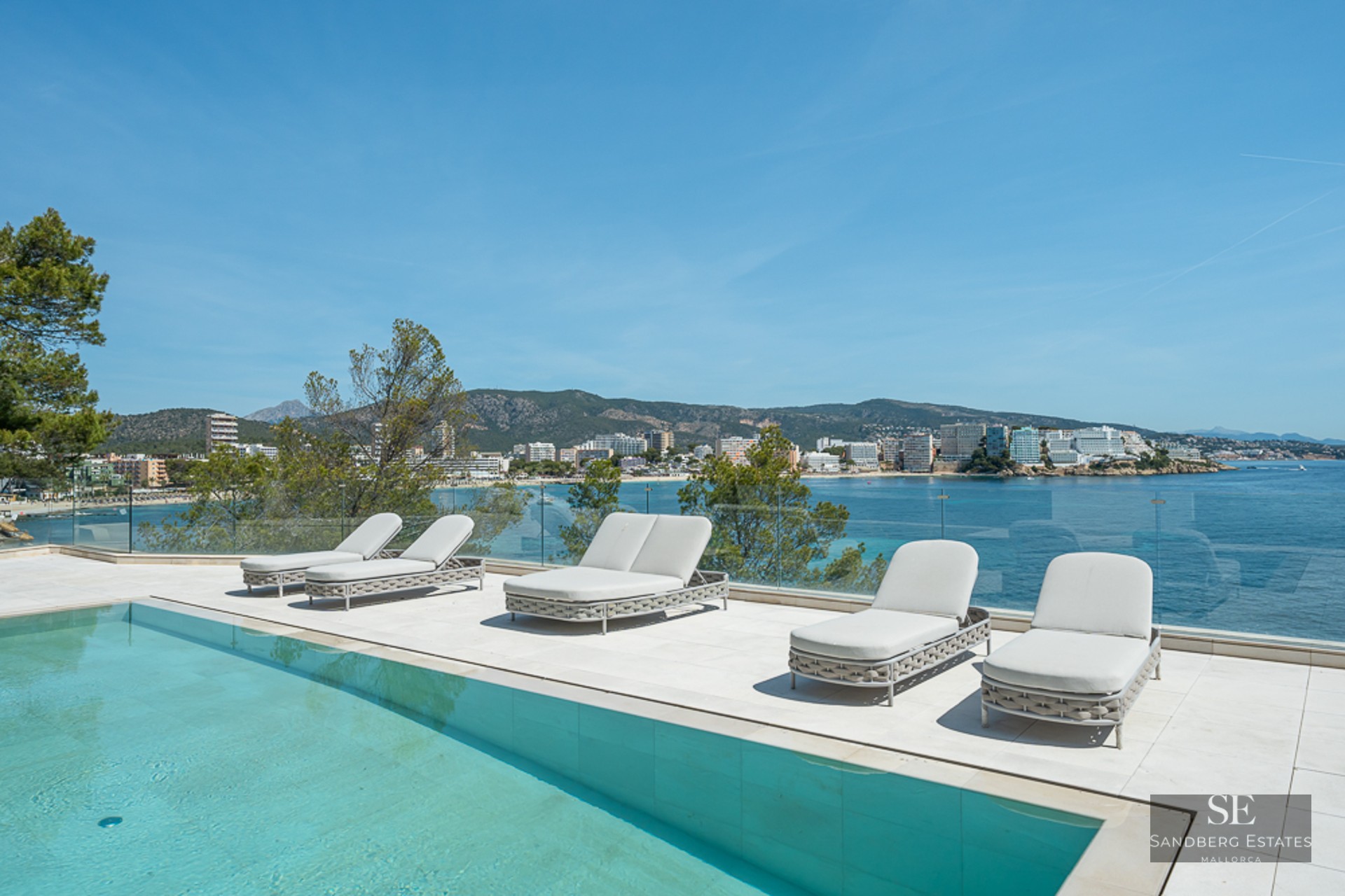 Modern white terrace with a turquoise pool, loungers, and a glass railing overlooking a scenic Mediterranean bay.