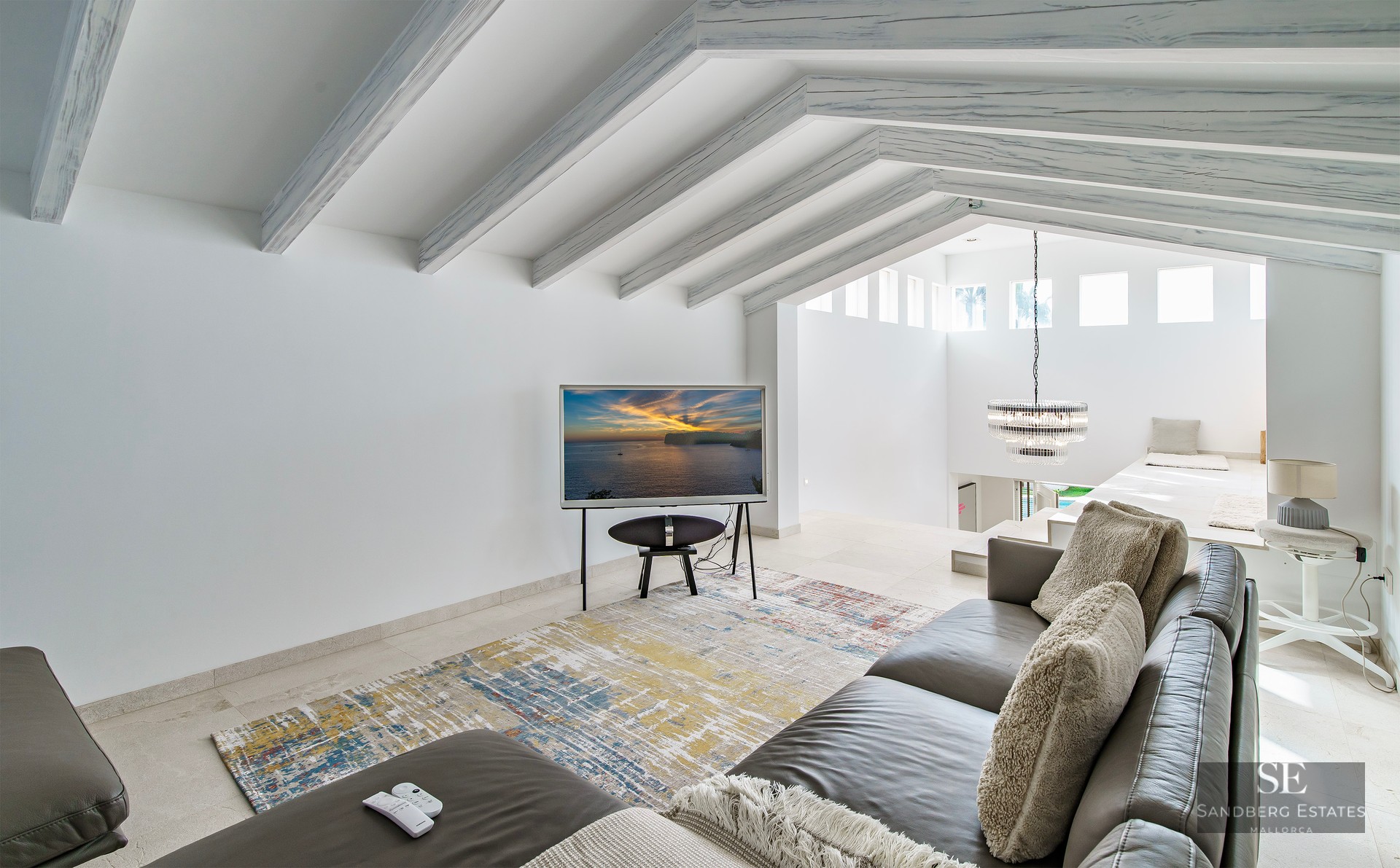 Bright living room featuring a grey leather sofa, abstract rug, and sloped ceiling with white-washed wooden beams.