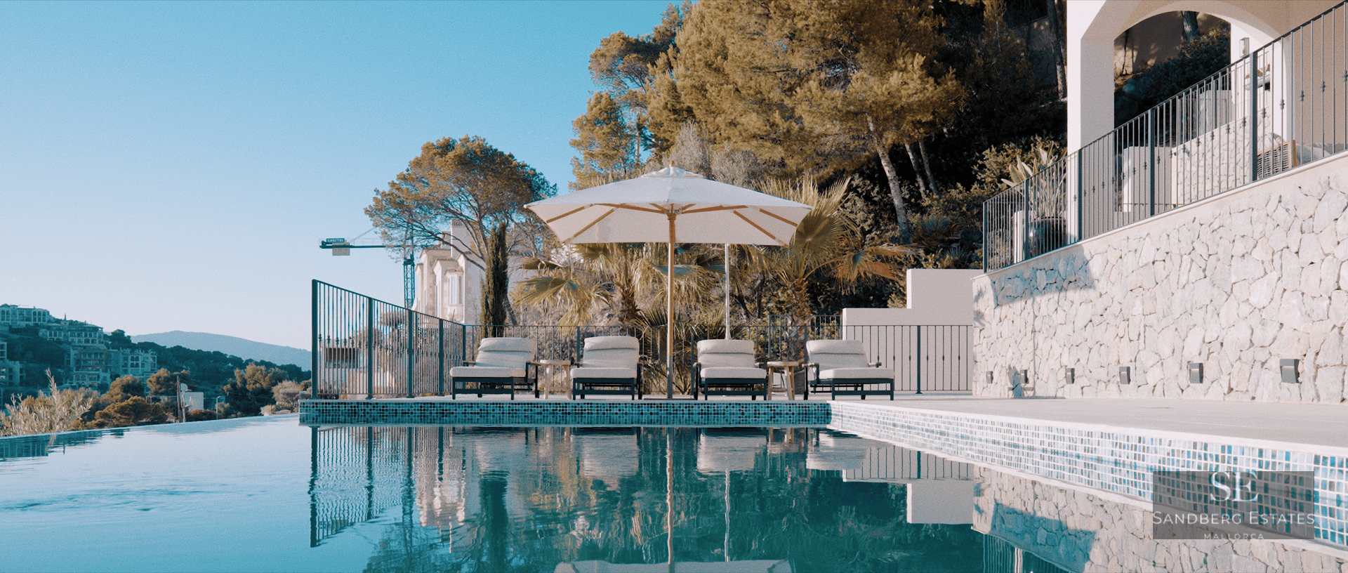 Infinity swimming pool with four sunbeds under a white umbrella, surrounded by stone walls and Mediterranean greenery.
