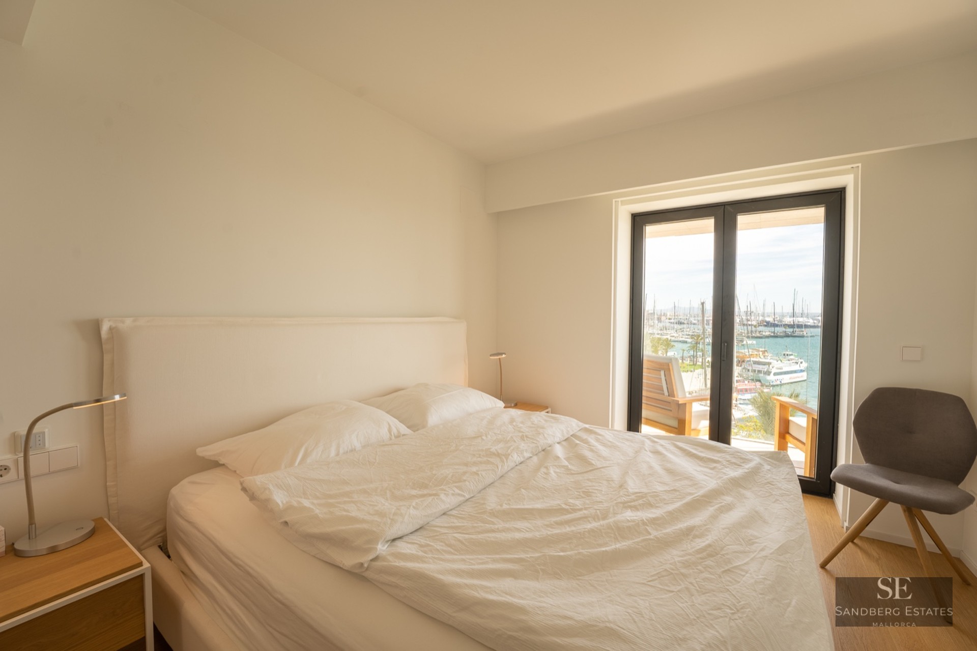 Bright minimalist bedroom with white linens, a grey chair, and large glass doors overlooking a marina with boats.