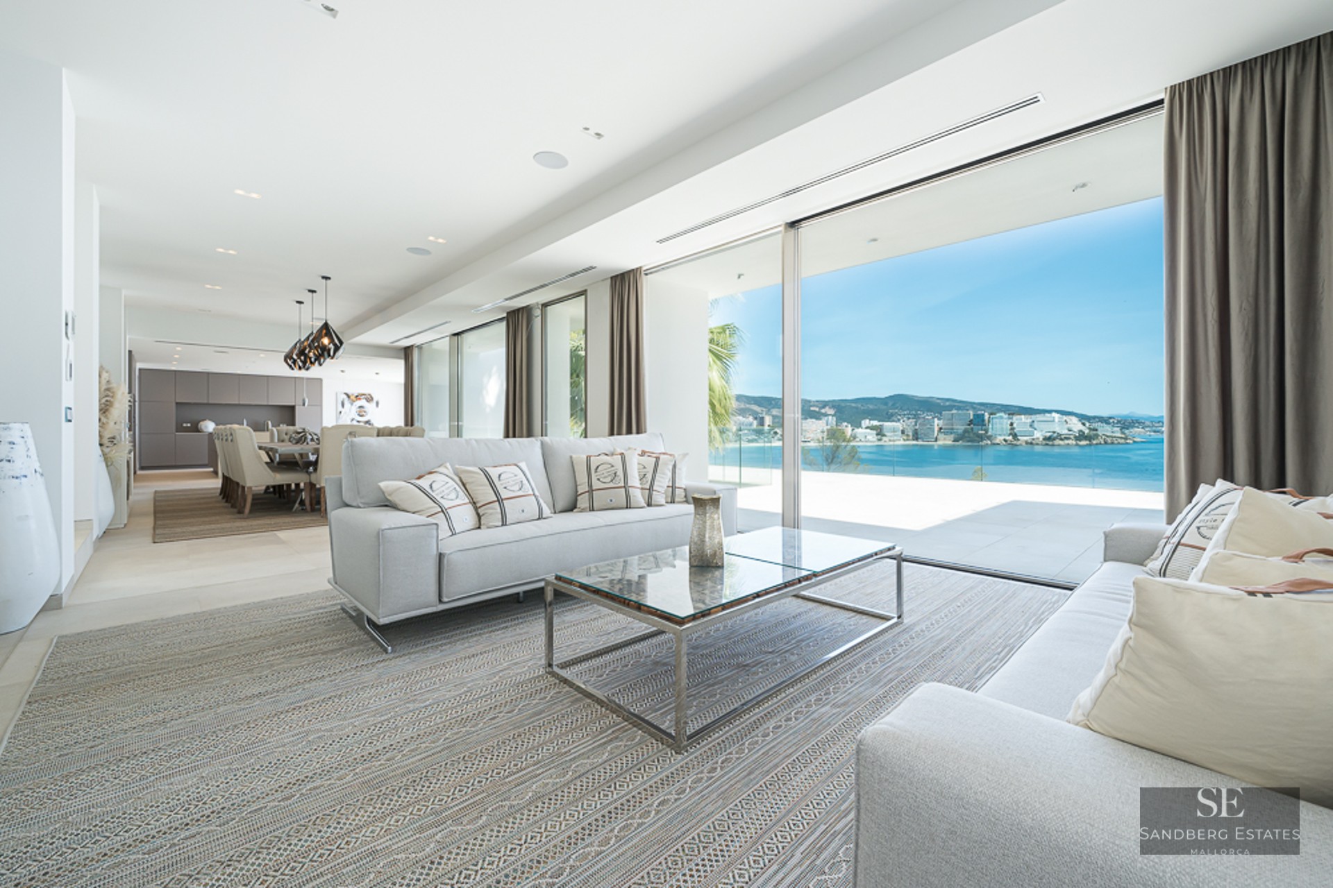 Modern living room with grey sofas and large glass doors opening to a panoramic view of the sea and coastline.