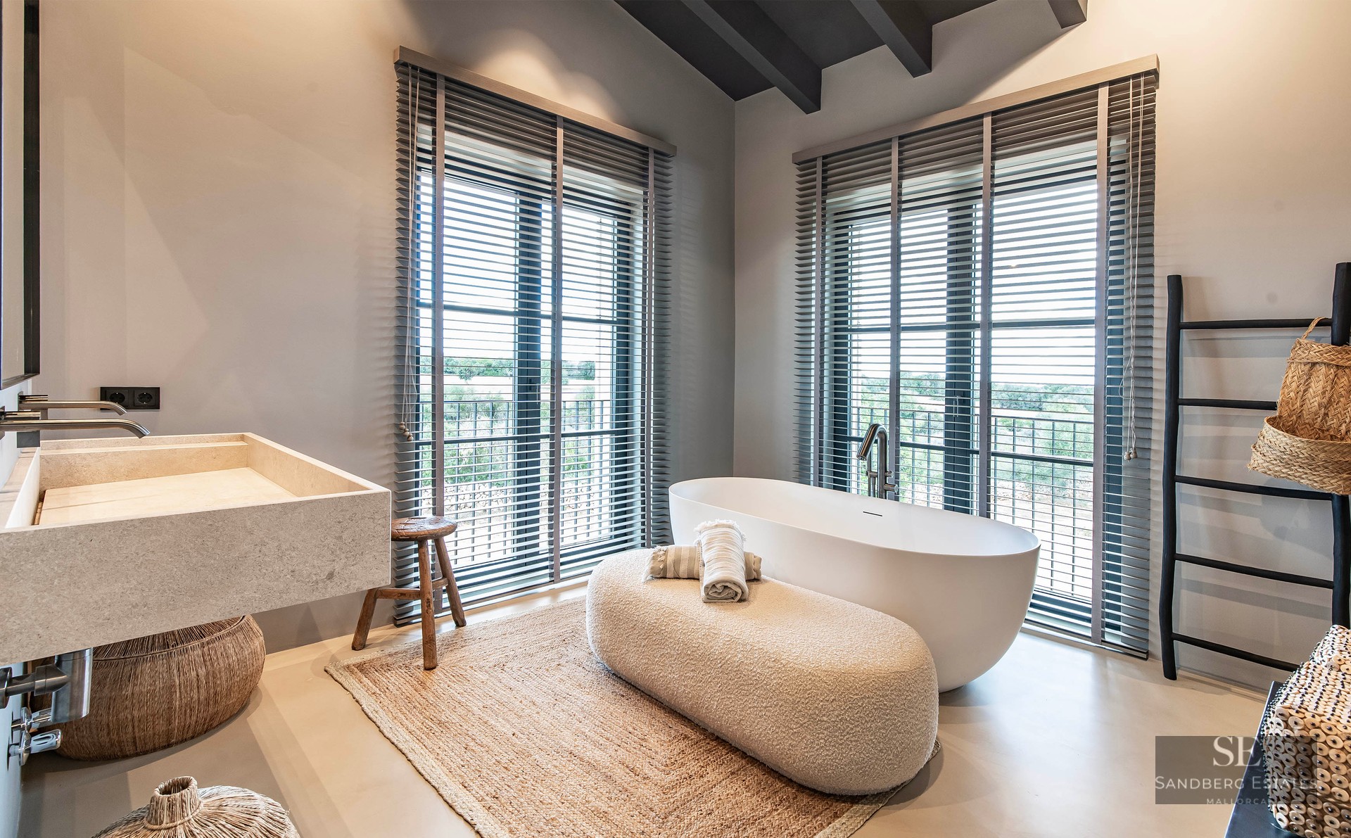 Modern master bathroom featuring a freestanding white tub, large stone sink, and windows with grey blinds.