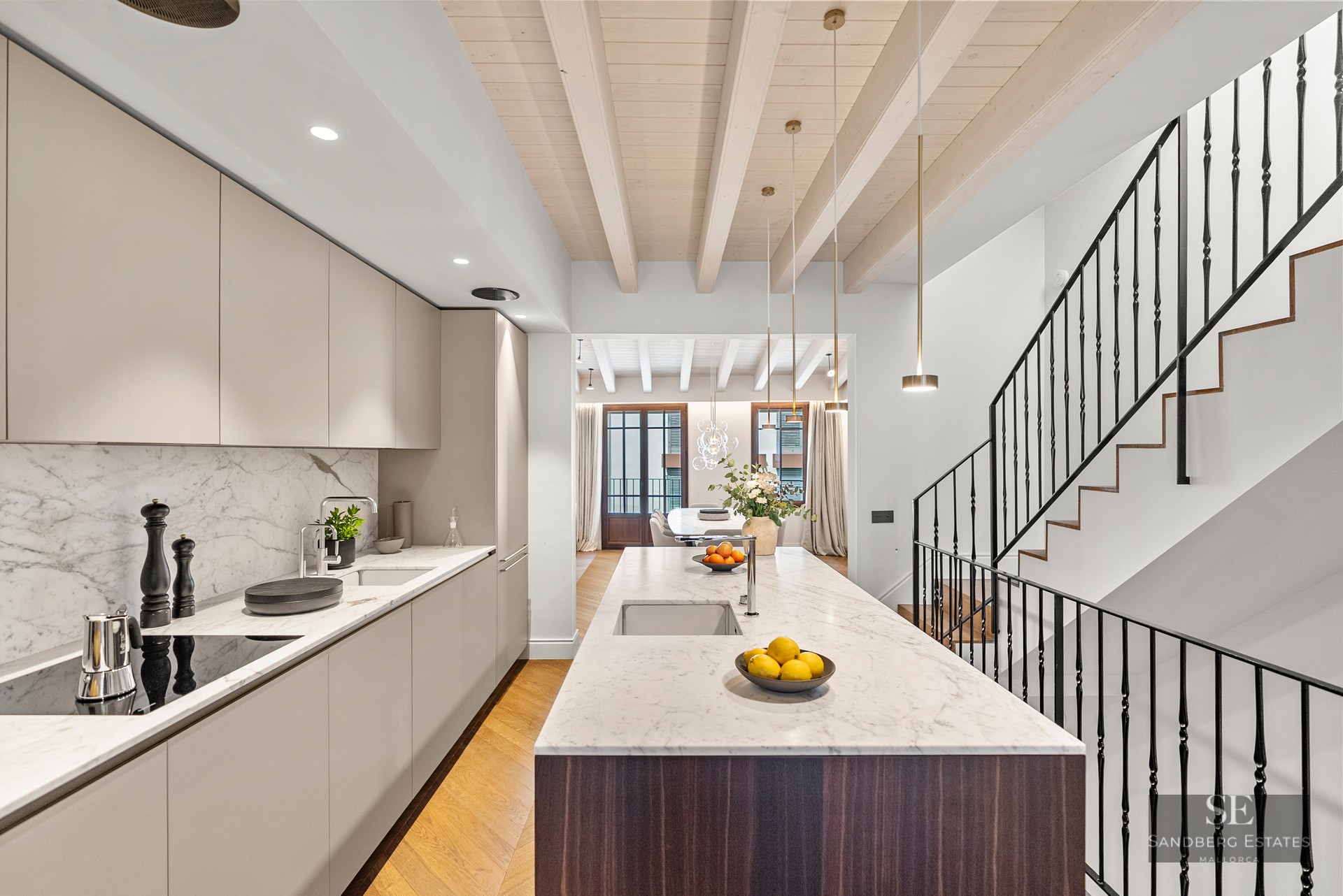 Contemporary kitchen featuring a large marble island, beige cabinetry, wooden ceiling beams, and a black staircase.