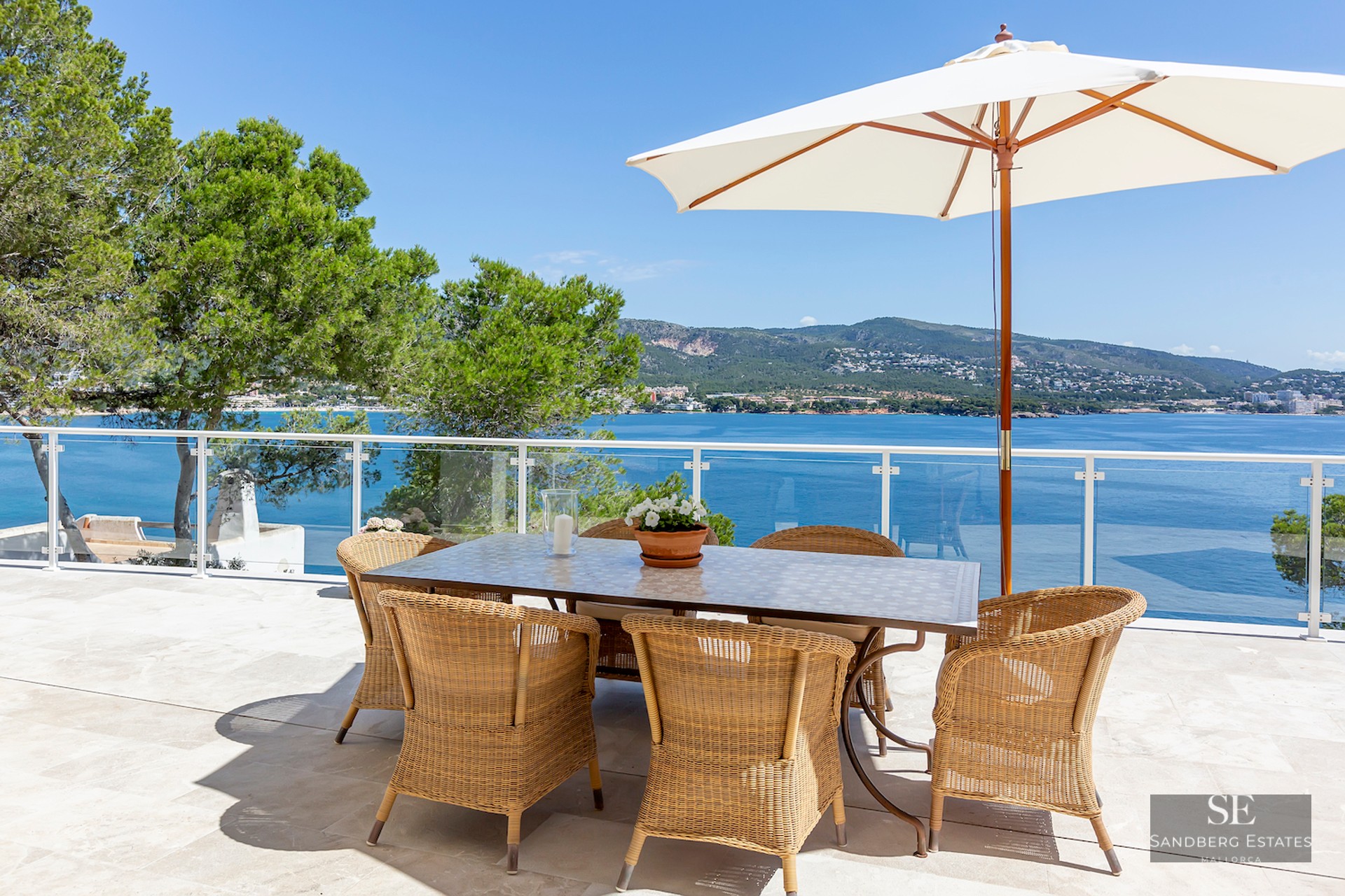 Outdoor dining area with wicker chairs and white umbrella on a stone terrace overlooking the blue sea and mountains.