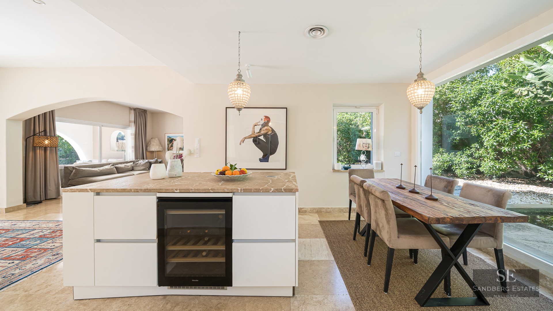 A modern white kitchen island with a wine fridge next to a rustic wooden dining table overlooking a lush garden.