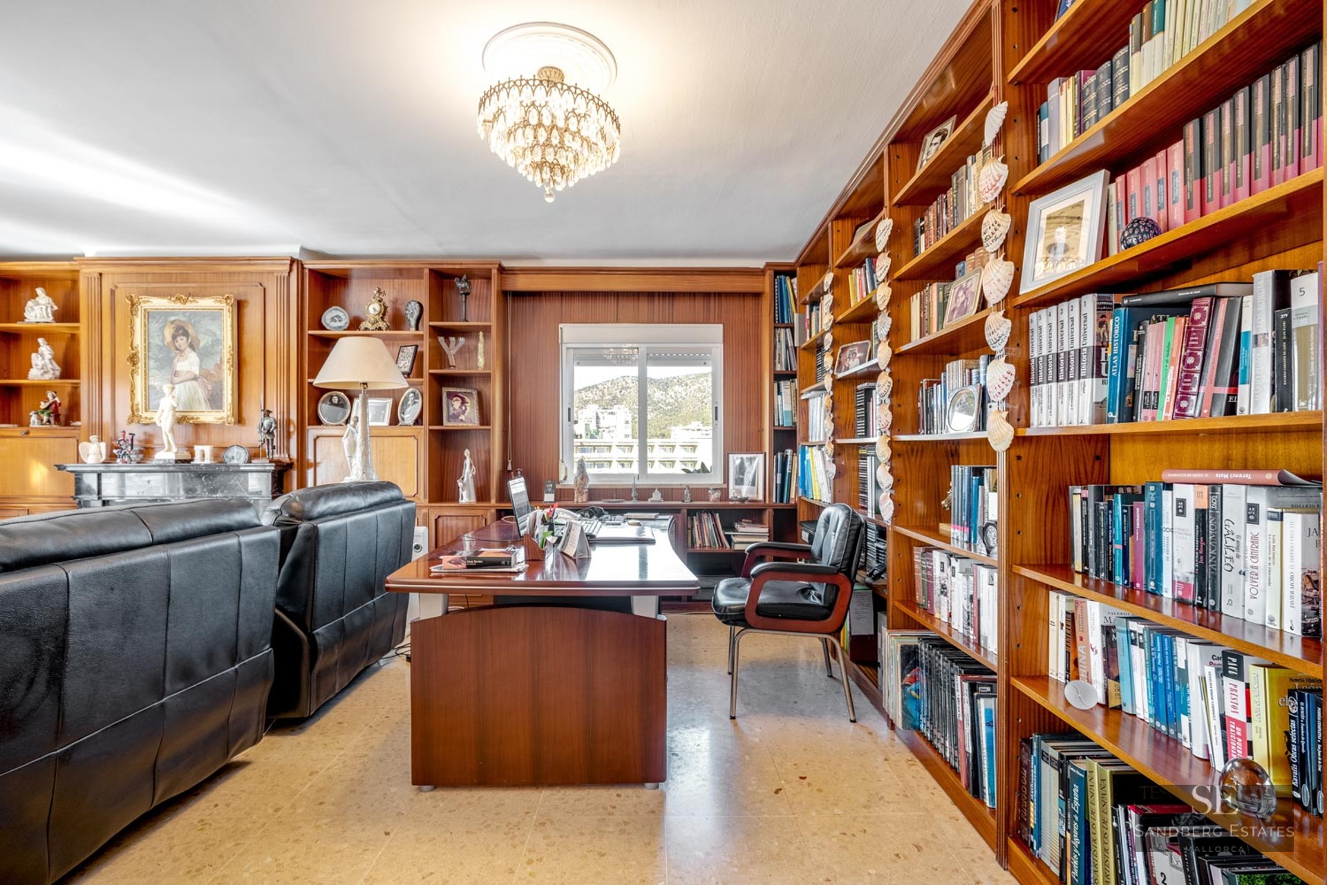Spacious home office featuring floor-to-ceiling wooden bookshelves, a large desk, leather seating, and a crystal chandelier.