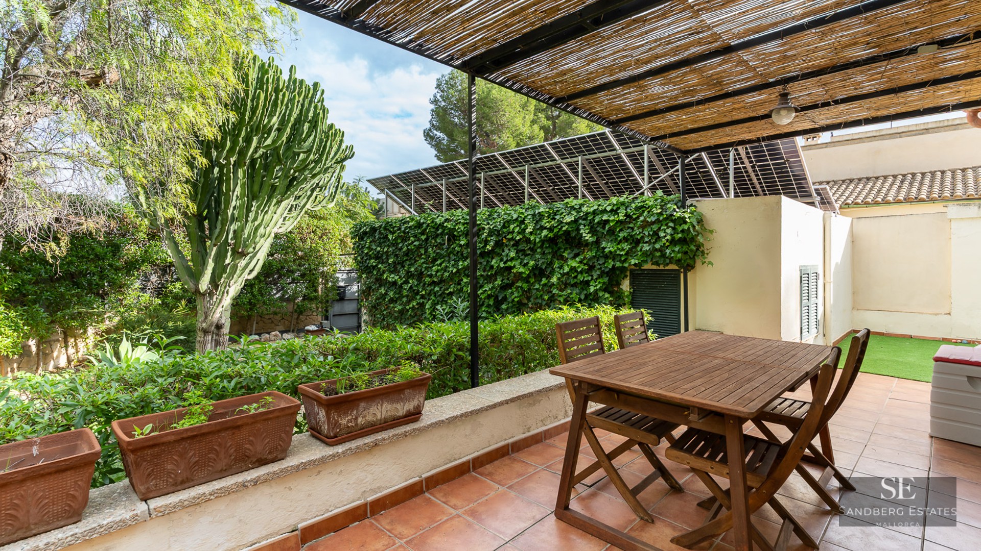 Wooden dining table under a bamboo pergola on a terracotta terrace surrounded by lush greenery and solar panels.
