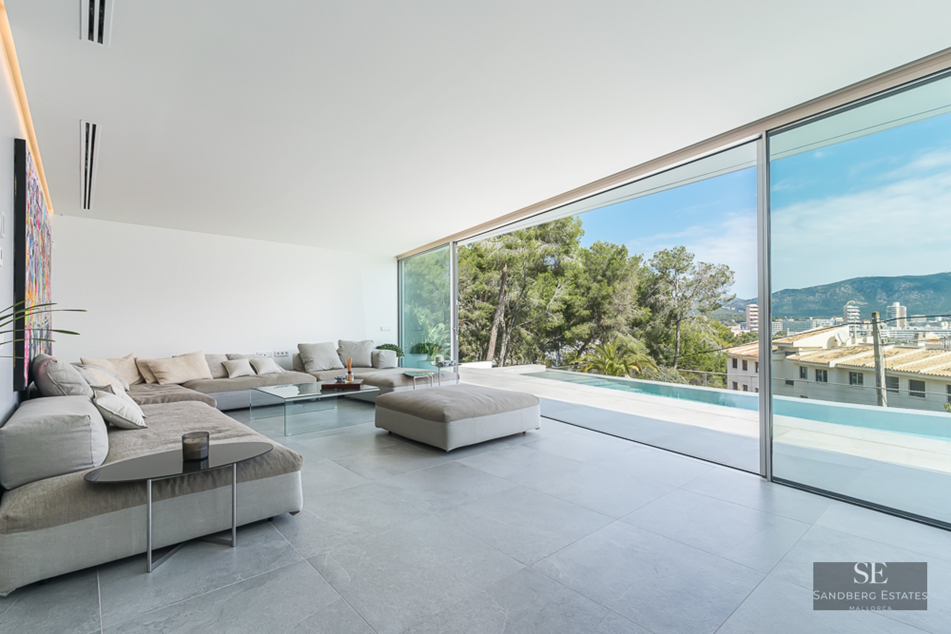 Spacious minimalist living room with grey sectional sofas and floor-to-ceiling glass doors overlooking a pool and mountains.
