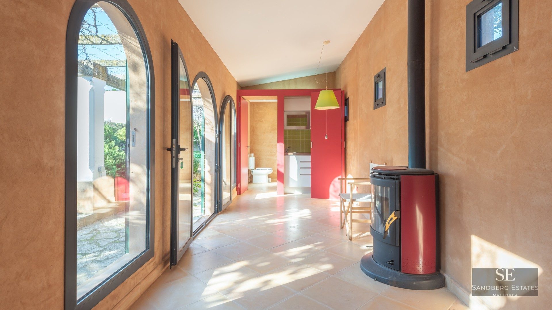 Sunlit room with terracotta walls, arched glass doors, a red wood-burning stove, and a view to an outdoor patio.