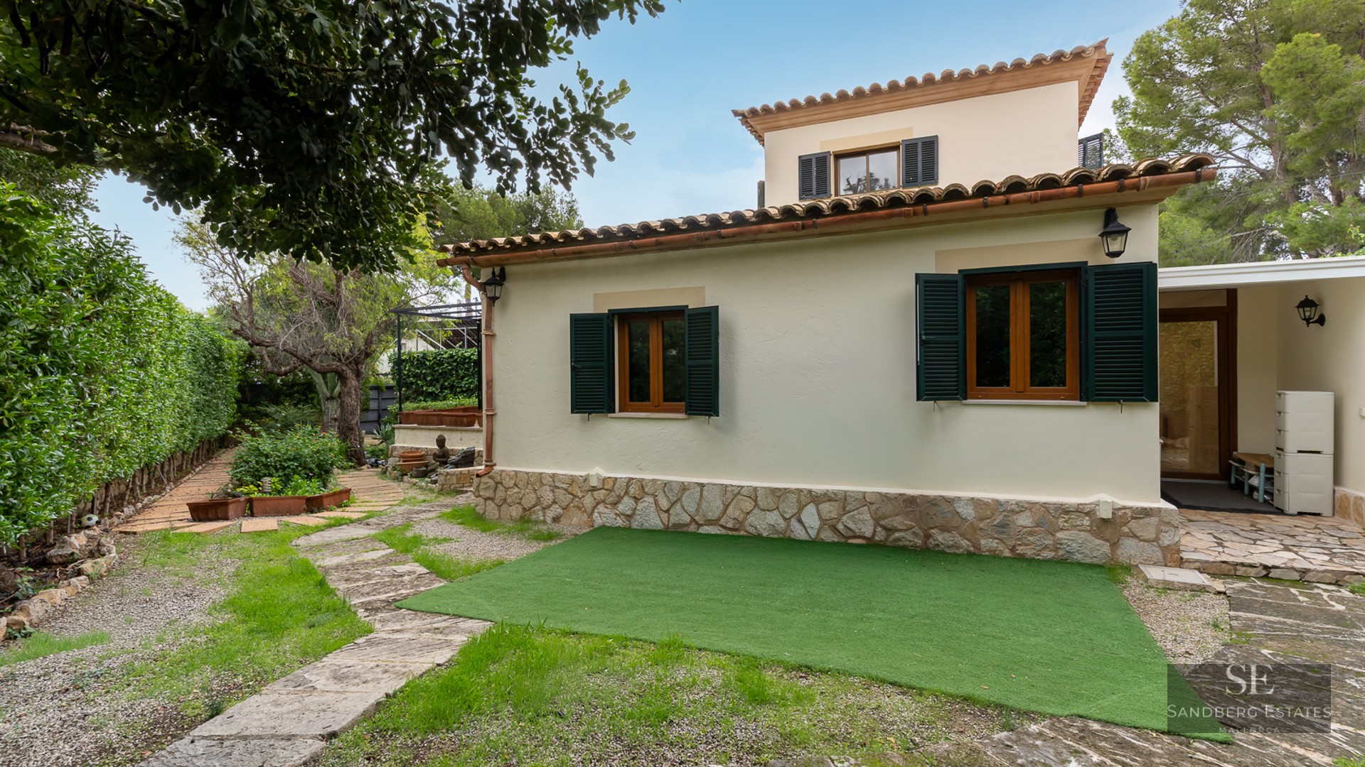 Cream stucco villa with green shutters, stone base, and a winding stone garden path under a clear blue sky.