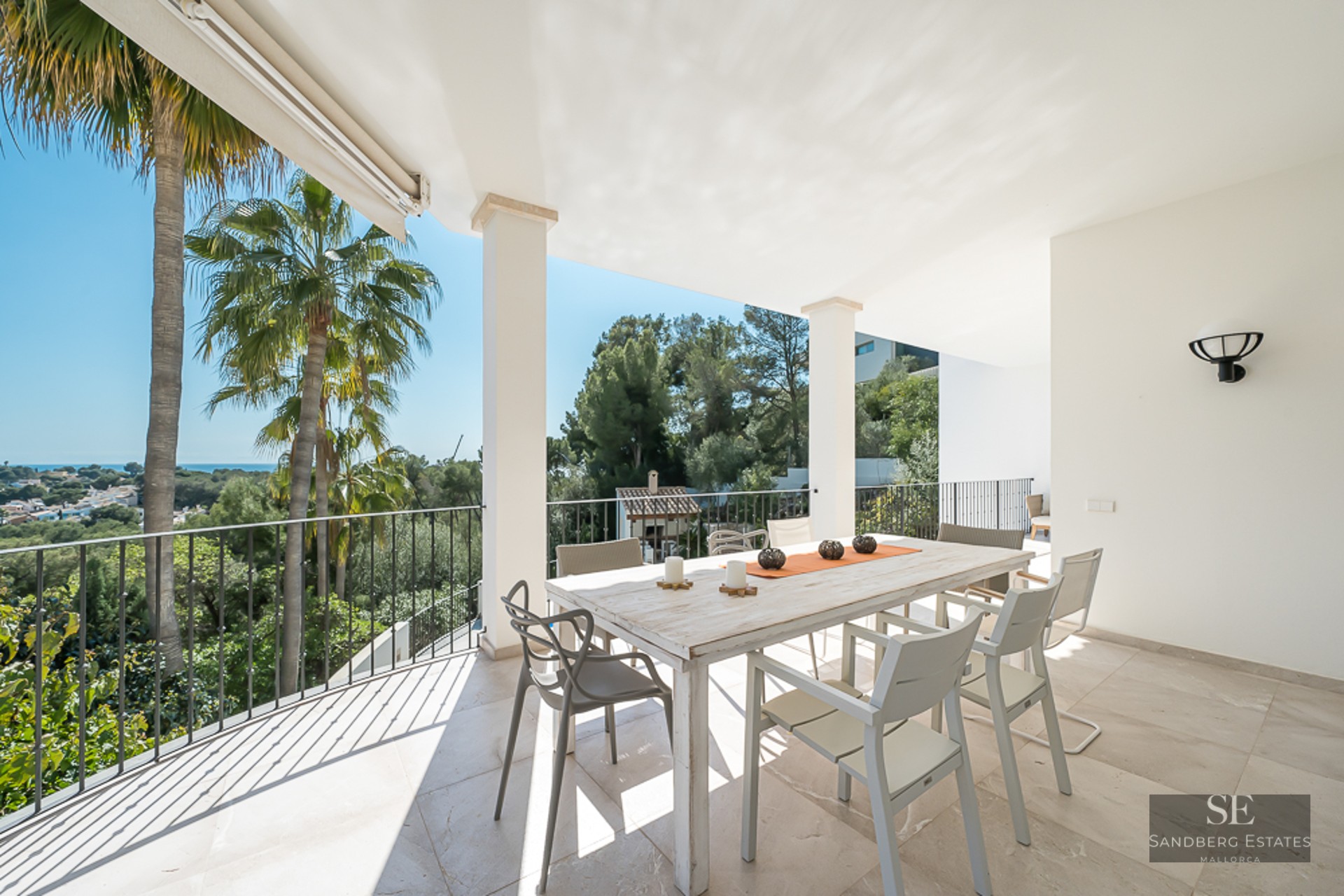 Terrasse blanche avec table à manger en bois, chaises modernes et vue sur les palmiers et la mer.