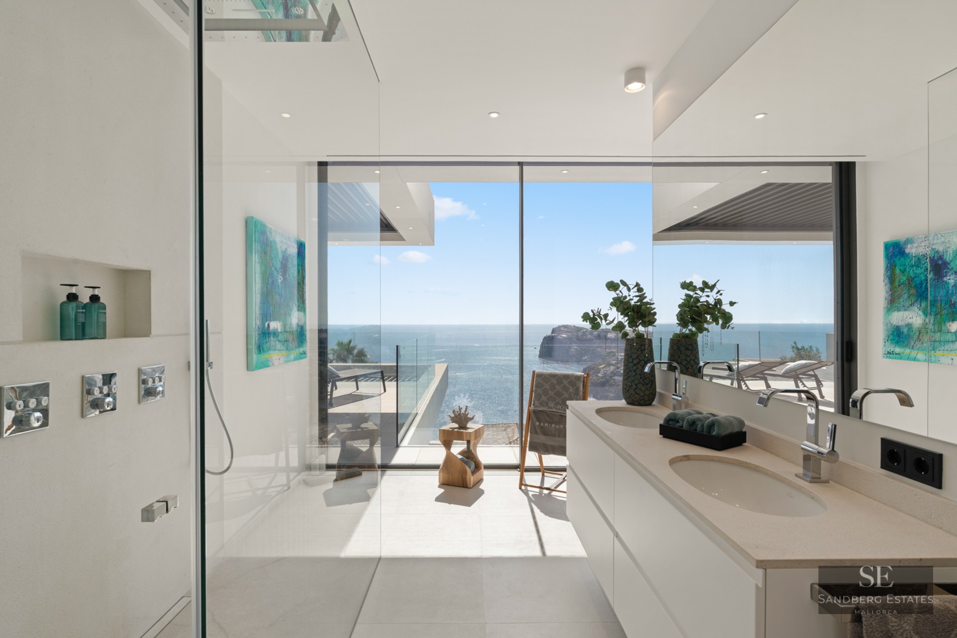 Modern white bathroom with glass walk-in shower and double vanity overlooking the ocean through floor-to-ceiling windows.
