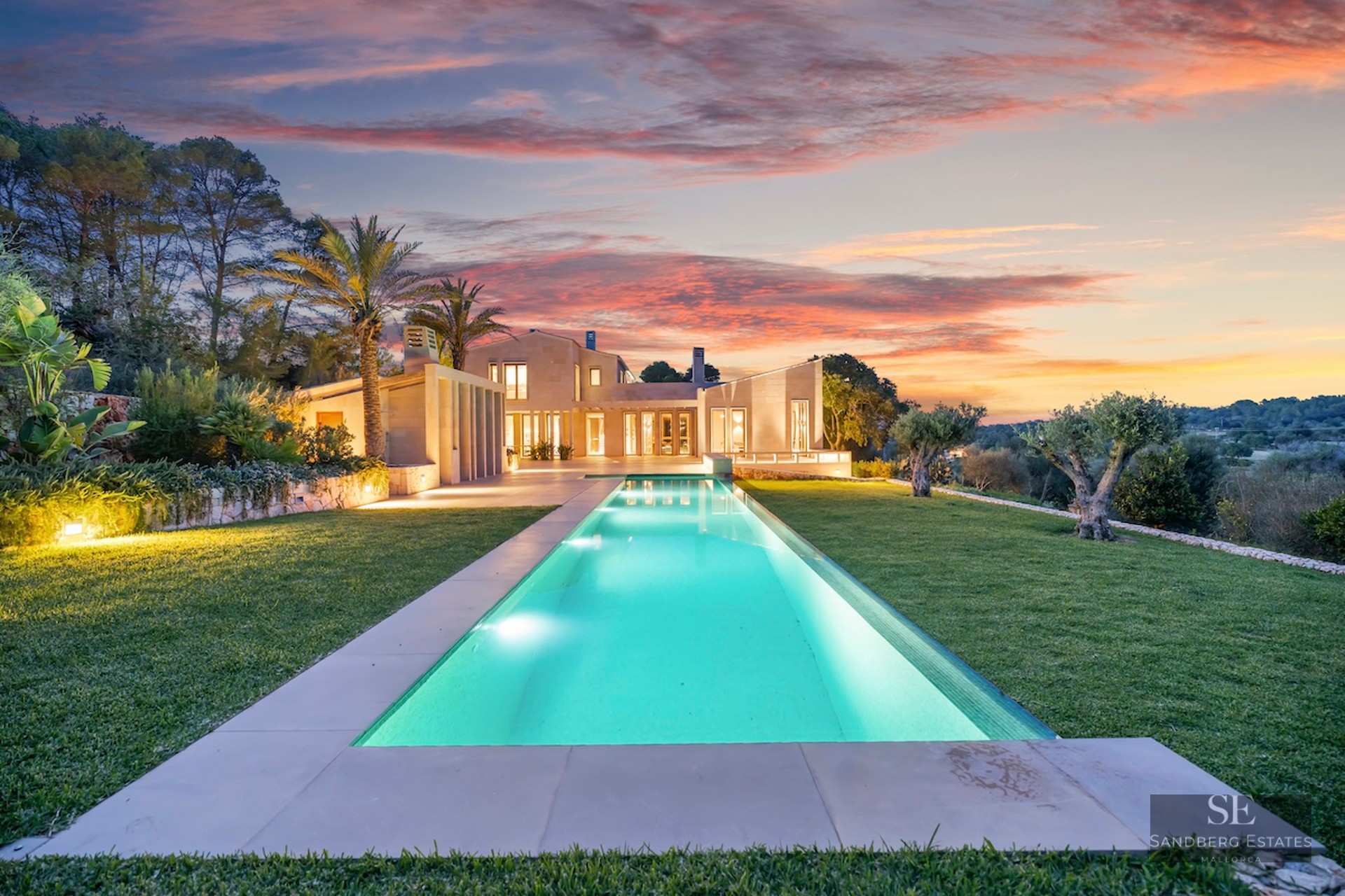 A long, illuminated rectangular pool leading to a modern stone villa under a vibrant pink and orange sunset sky.