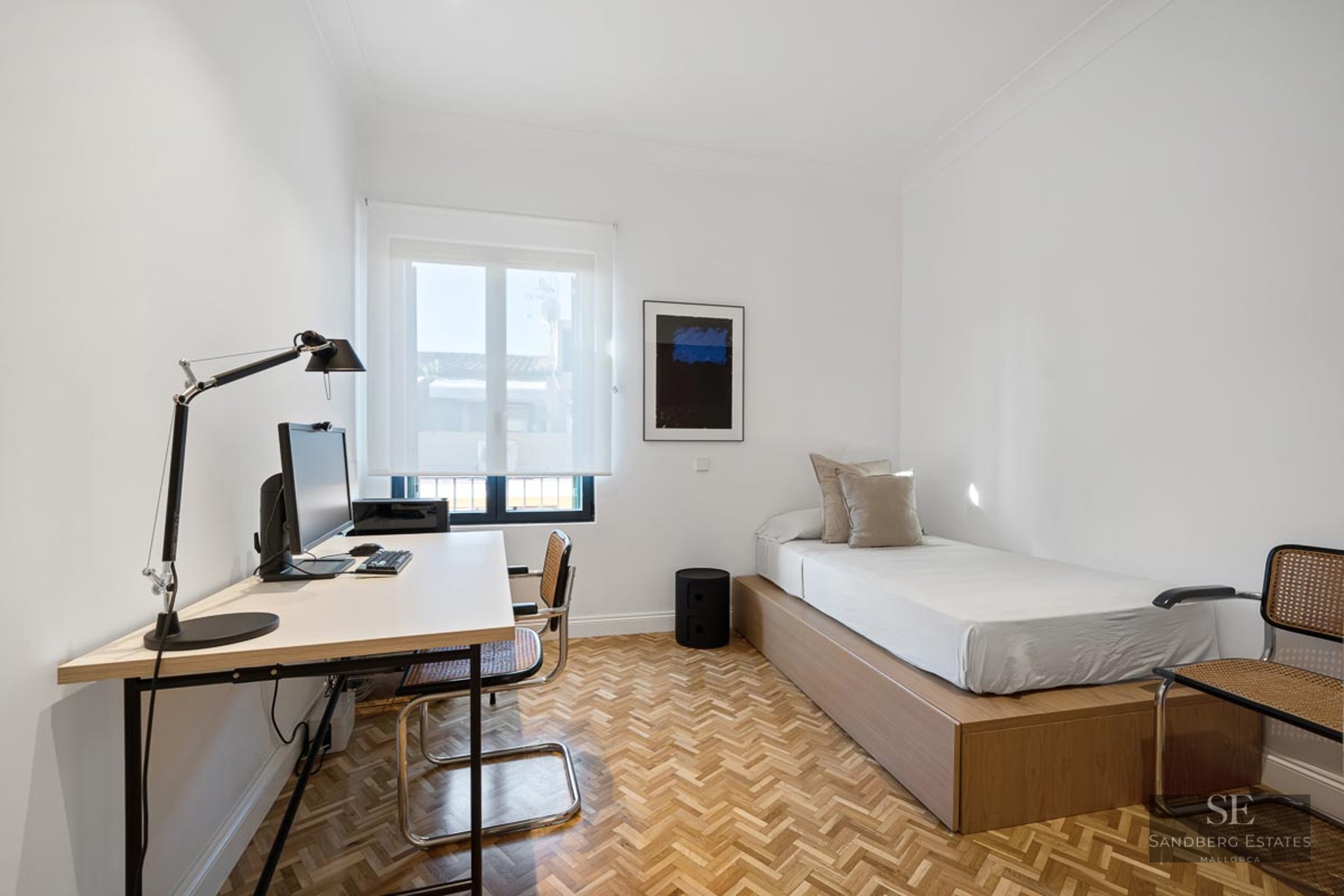 Bright minimalist room featuring a single bed, herringbone parquet floors, and a desk with a computer.
