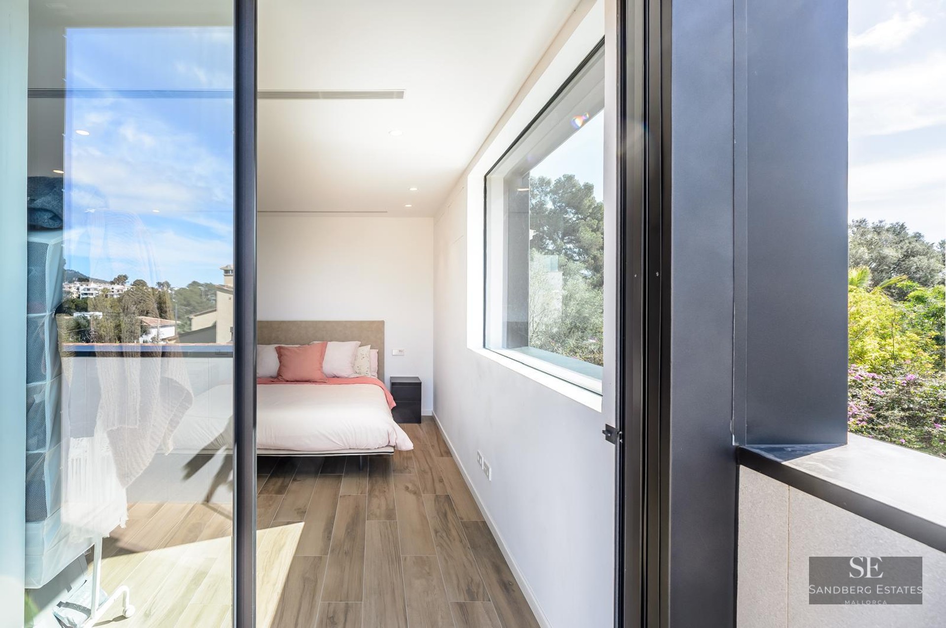 Modern bedroom with wood floors, a bed with pink pillows, and large windows overlooking green trees.