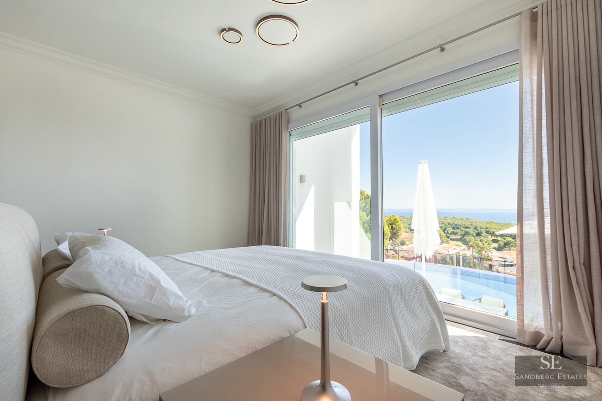 Modern bedroom with white bedding, large windows overlooking a private infinity pool and distant sea view.