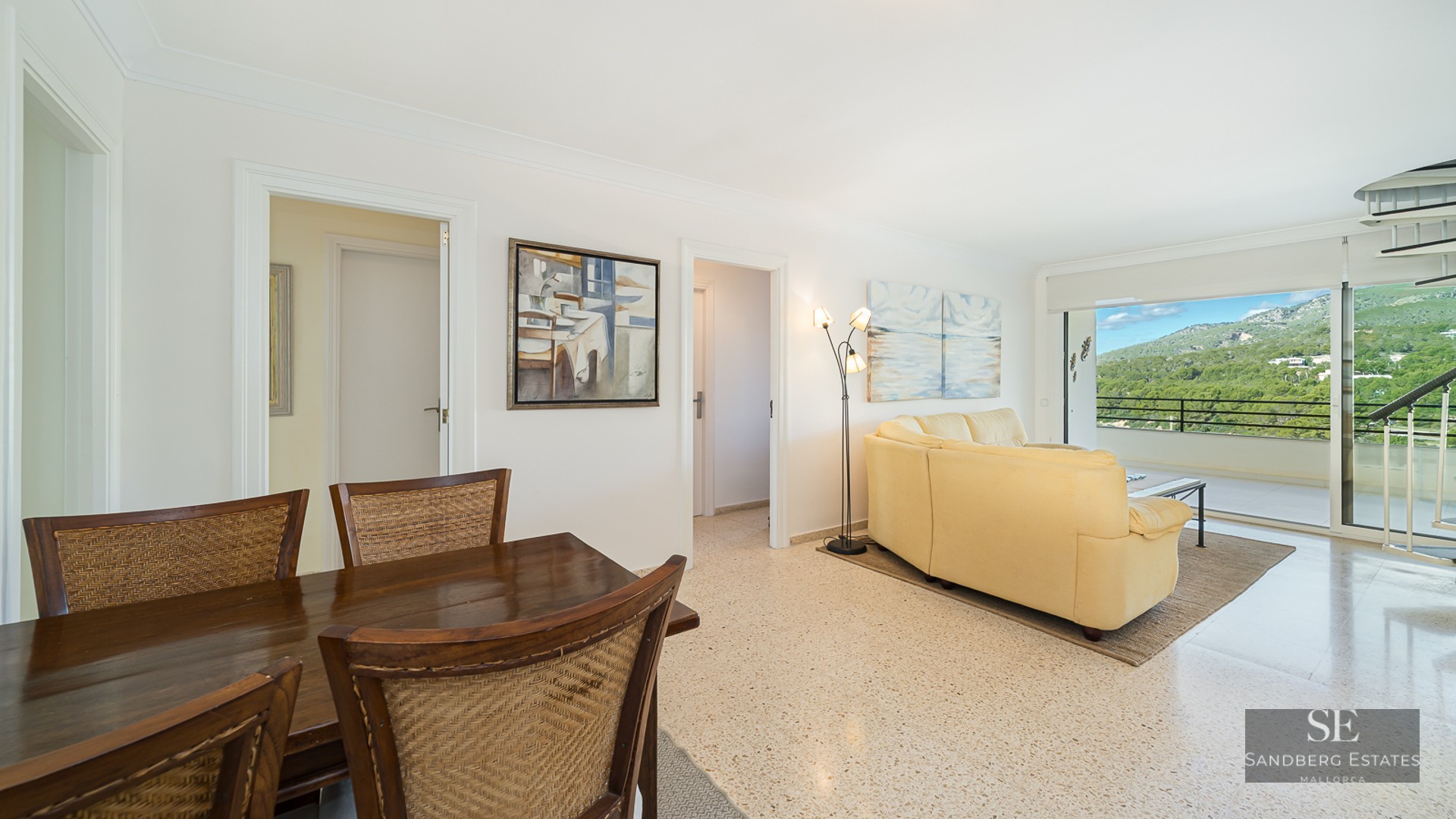 Open living space with a cream leather sofa, wooden dining set, and large glass doors revealing a mountain landscape.