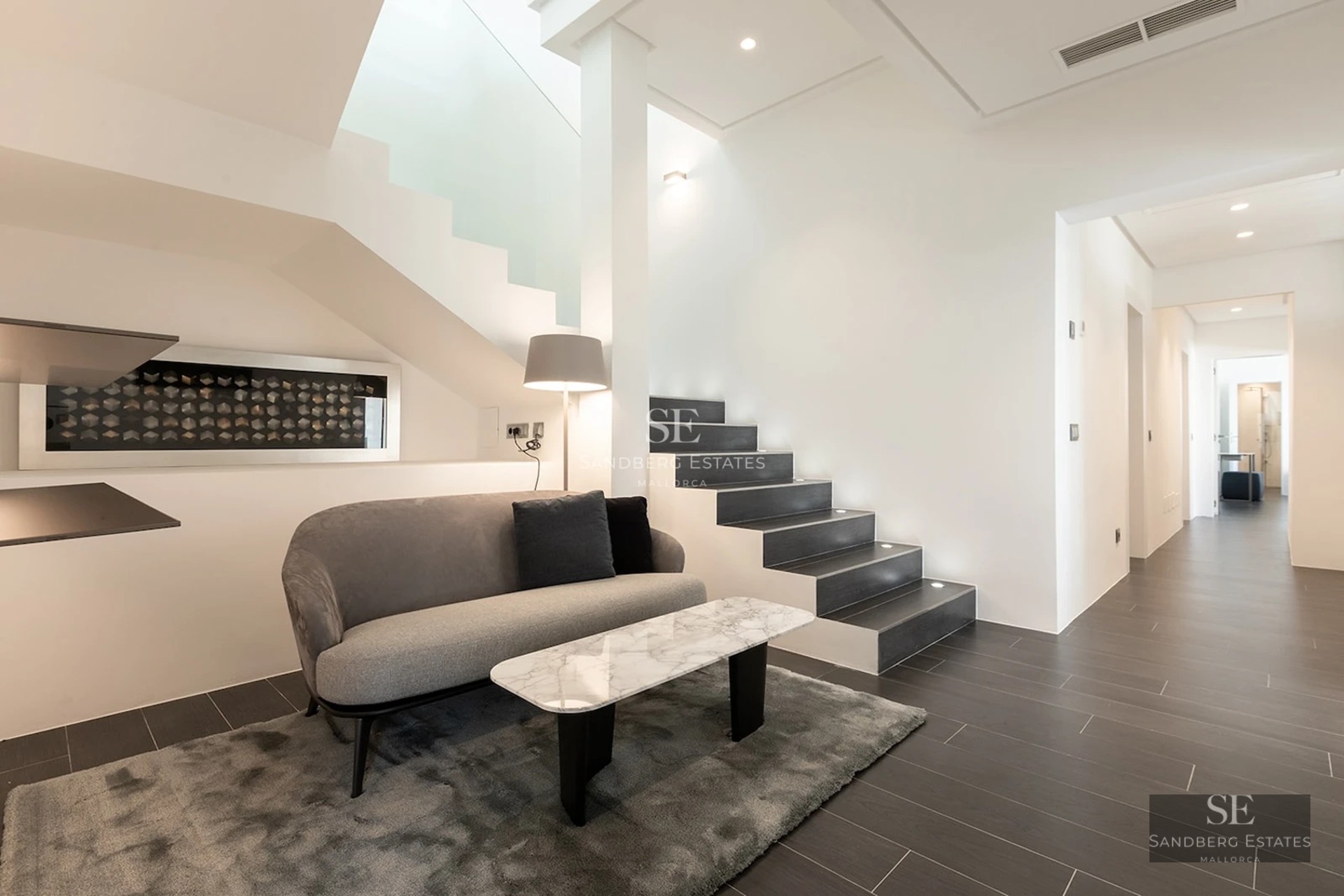 Modern minimalist lounge area with a grey sofa, marble coffee table, and a sleek dark staircase against white walls.
