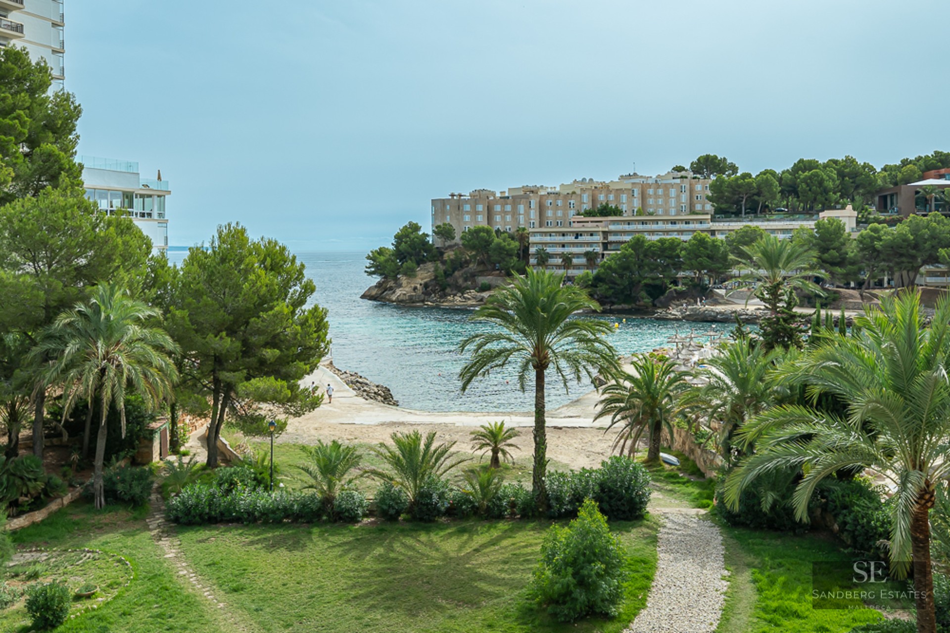 A lush garden with palm trees and a stone path overlooking a turquoise Mediterranean bay and coastal buildings.