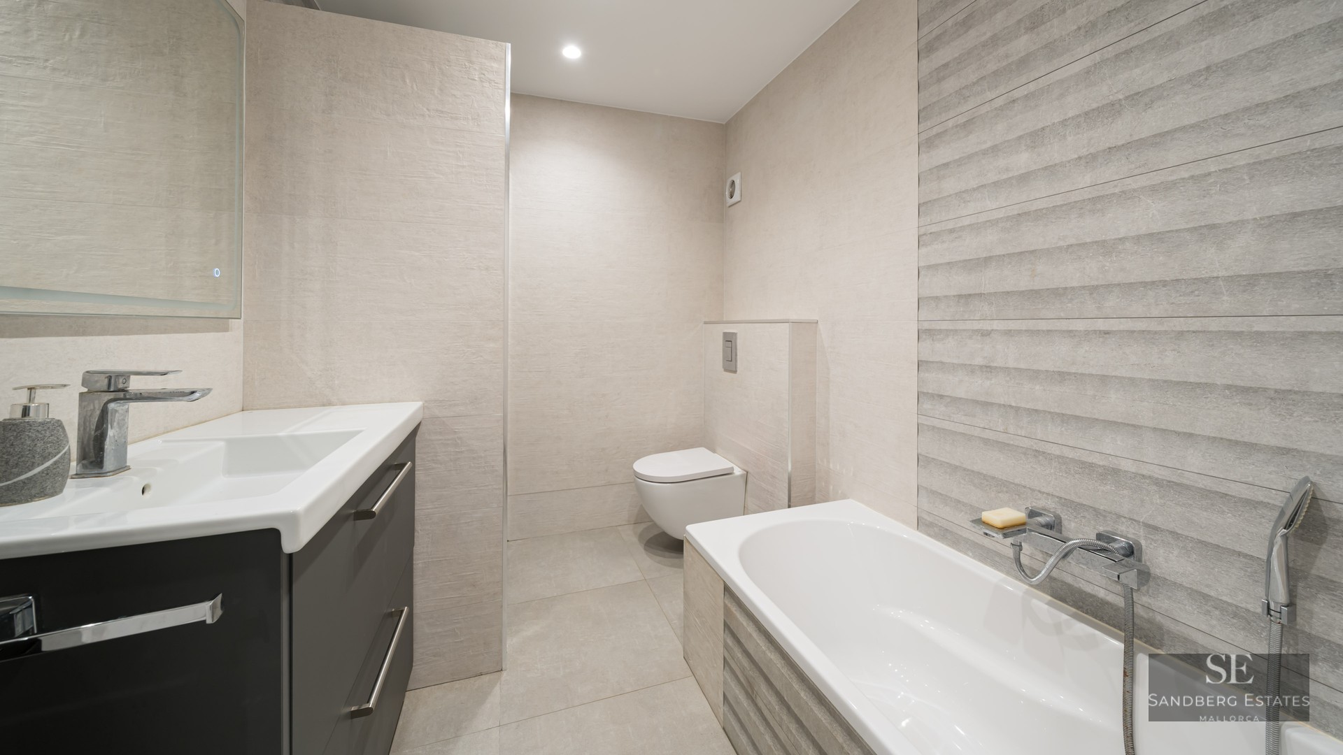 Salle de bain moderne avec baignoire blanche, WC suspendu et meuble gris sur fond de carrelage beige texturé.