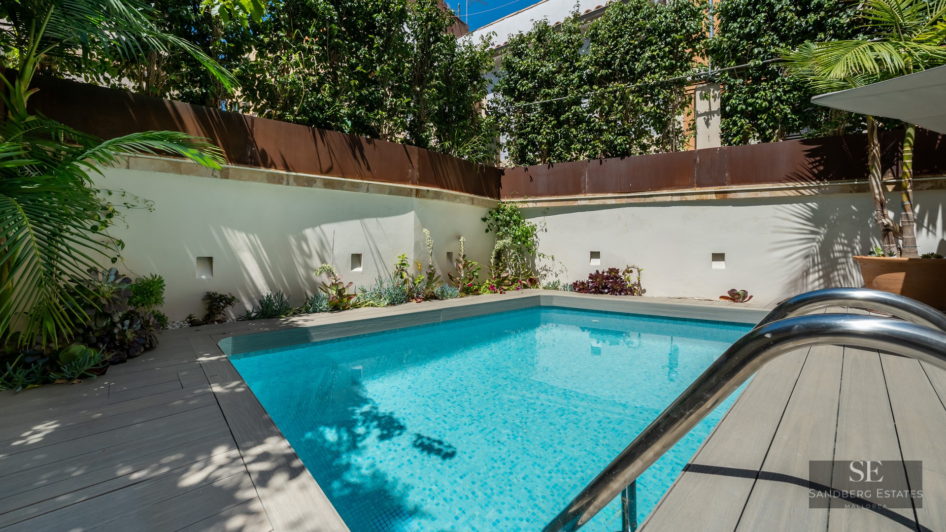 Small turquoise swimming pool with grey wooden decking surrounded by white walls and lush green plants.