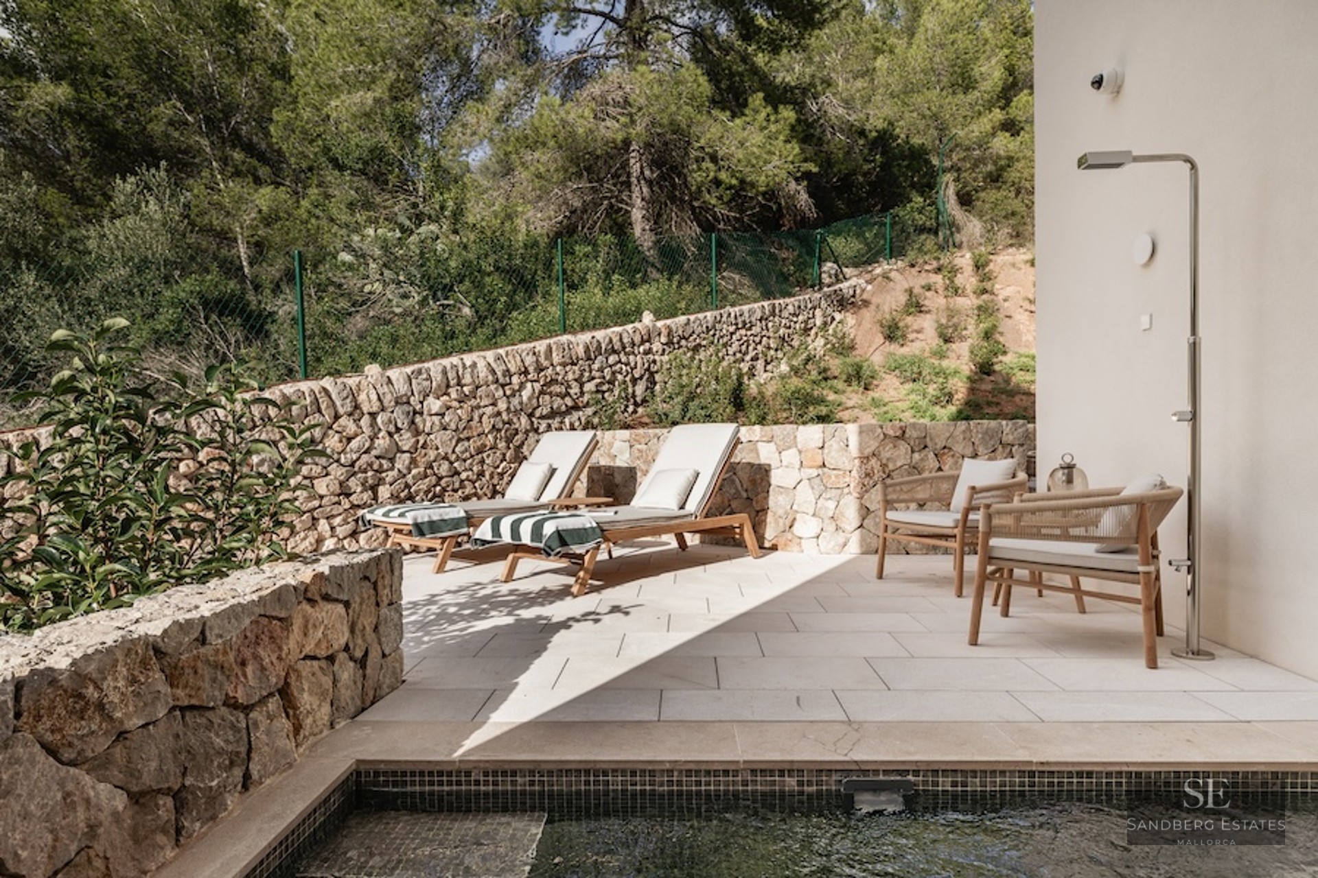 Modern stone terrace featuring designer sun loungers, a metallic outdoor shower, and views of a lush forest.