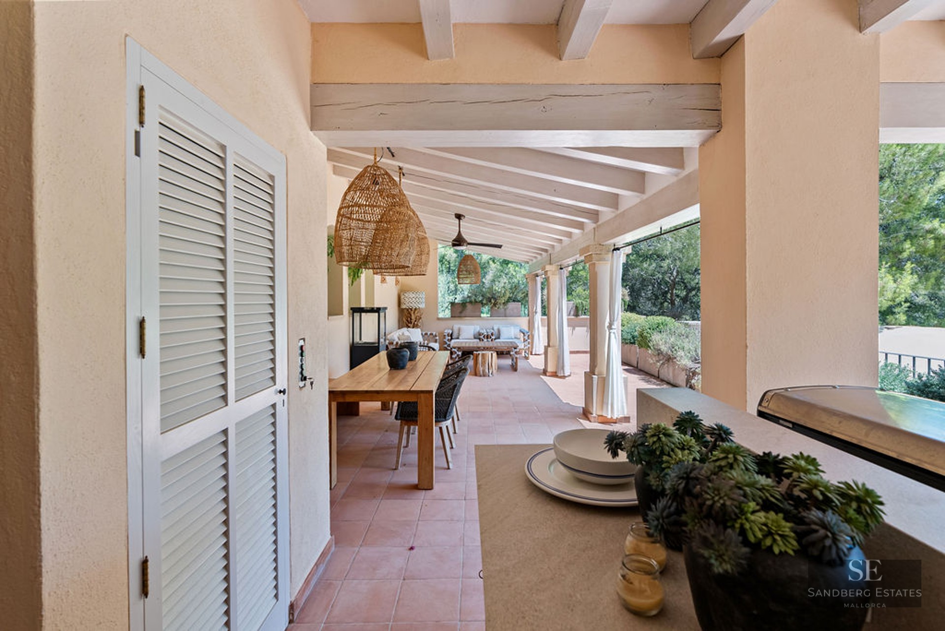 Large covered terrace with terracotta floors, exposed beams, a long wooden dining table, and wicker pendant lights.
