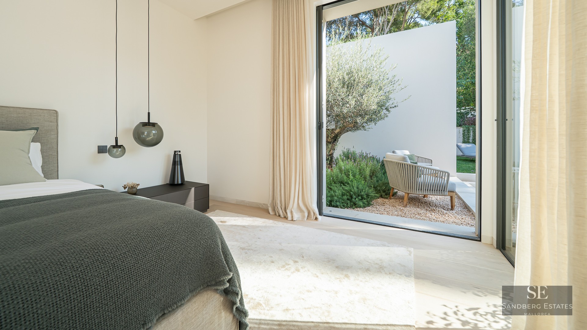 Modern bedroom with a large bed, designer pendant lights, and sliding glass doors opening to a sunlit garden with an olive tree.
